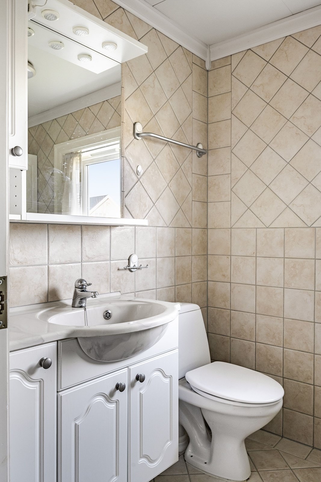 Baderom i 1. etasje med overflater, installasjoner og komponenter fra ca. 2005. Lys baderomsinnredning med heldekkende servant, speil med belysning og vegghengt skap over. Galleribilde