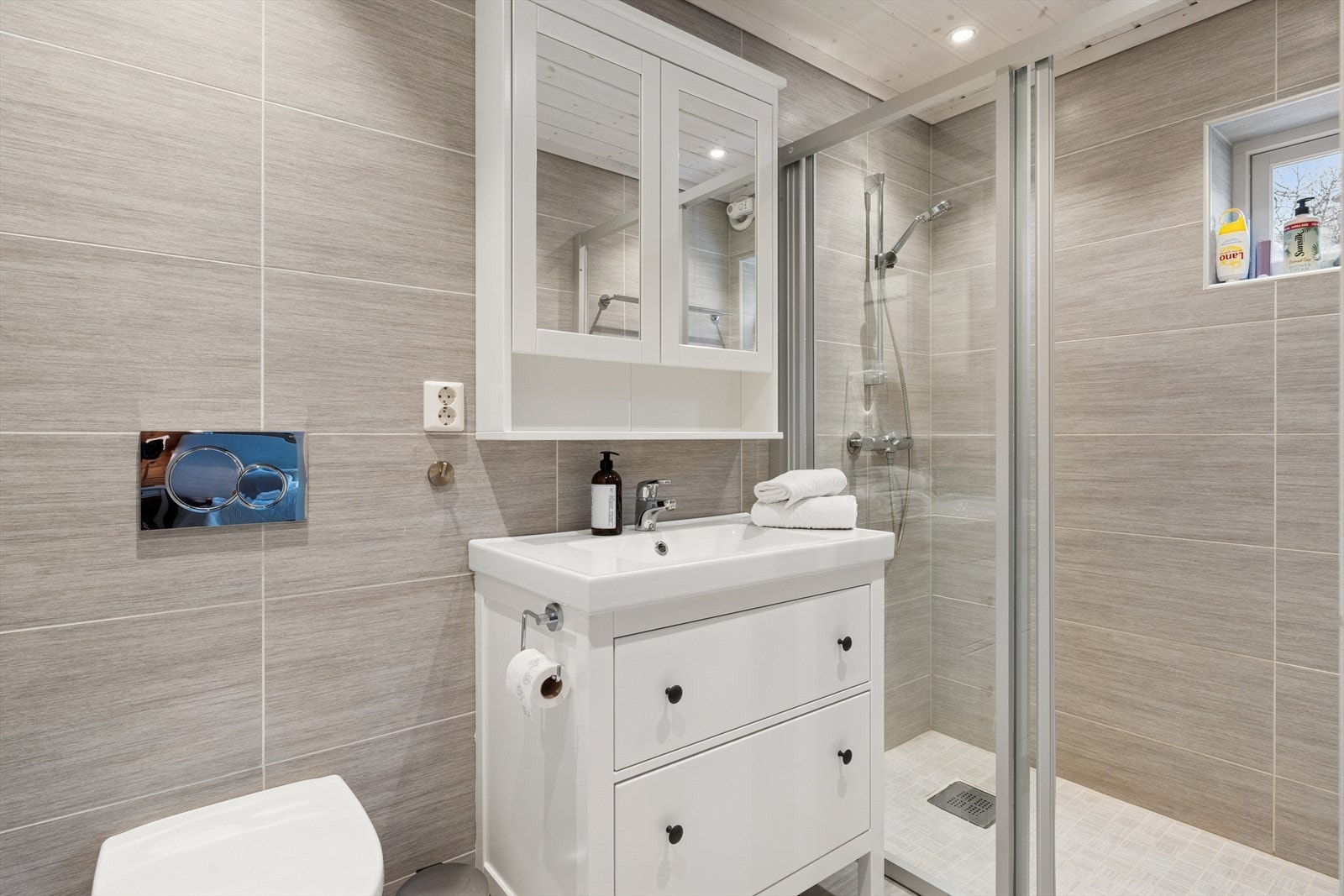 Flislagte vegger og elektrisk styrt vifte gir effektiv ventilasjon på badet. Dusjnisje med skyvedør gir praktisk utnyttelse av rommet. Galleribilde