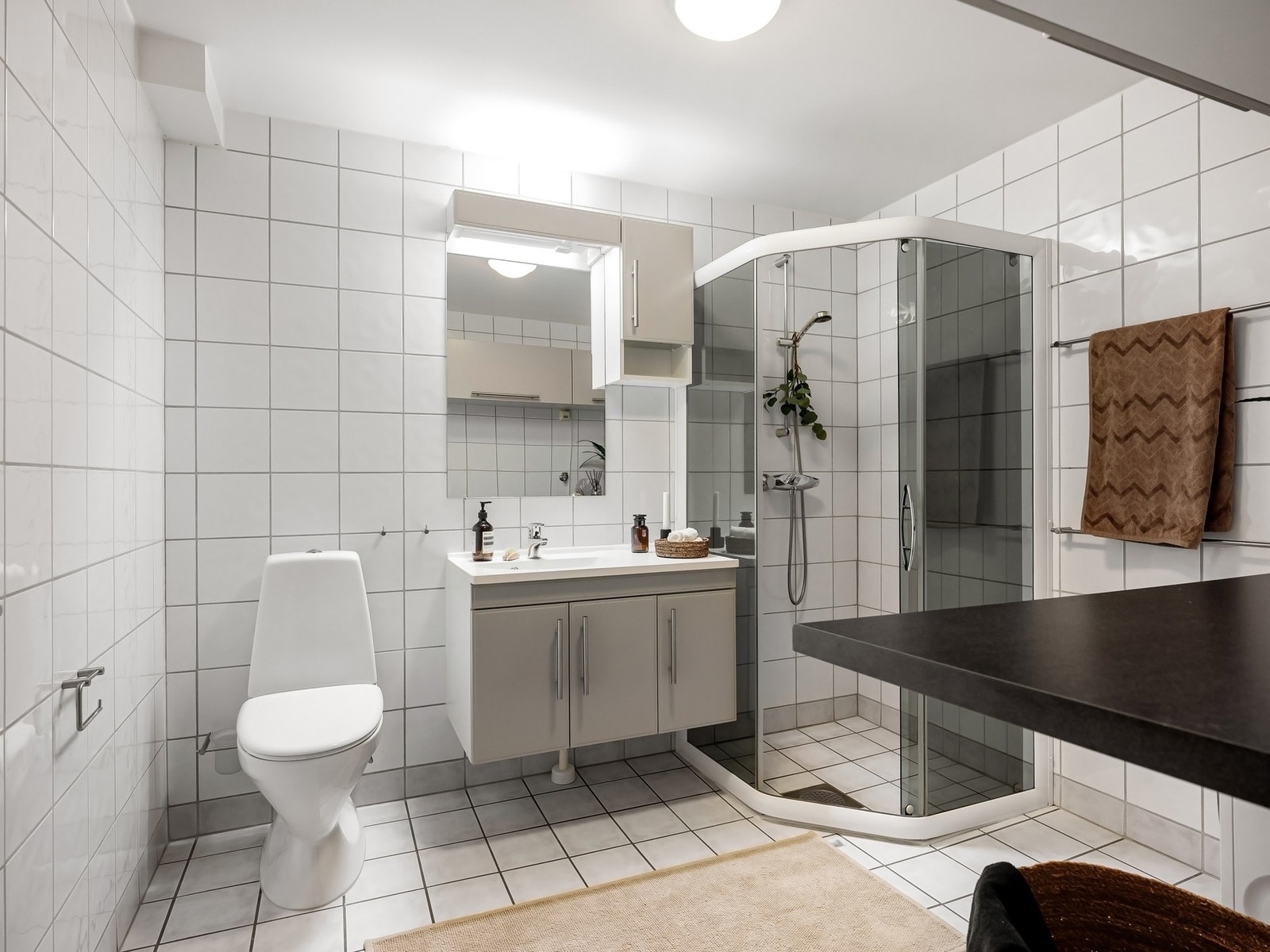 Rommet er innredet med gulvstående toalett, servant med ettgreps armatur og romslig skap under. Over servanten finner du speil med lyslist og innebygd stikkontakt. Dusjsonen består av et elegant dusjhørne i stål og glass, og det er i tillegg opplegg for v Galleribilde