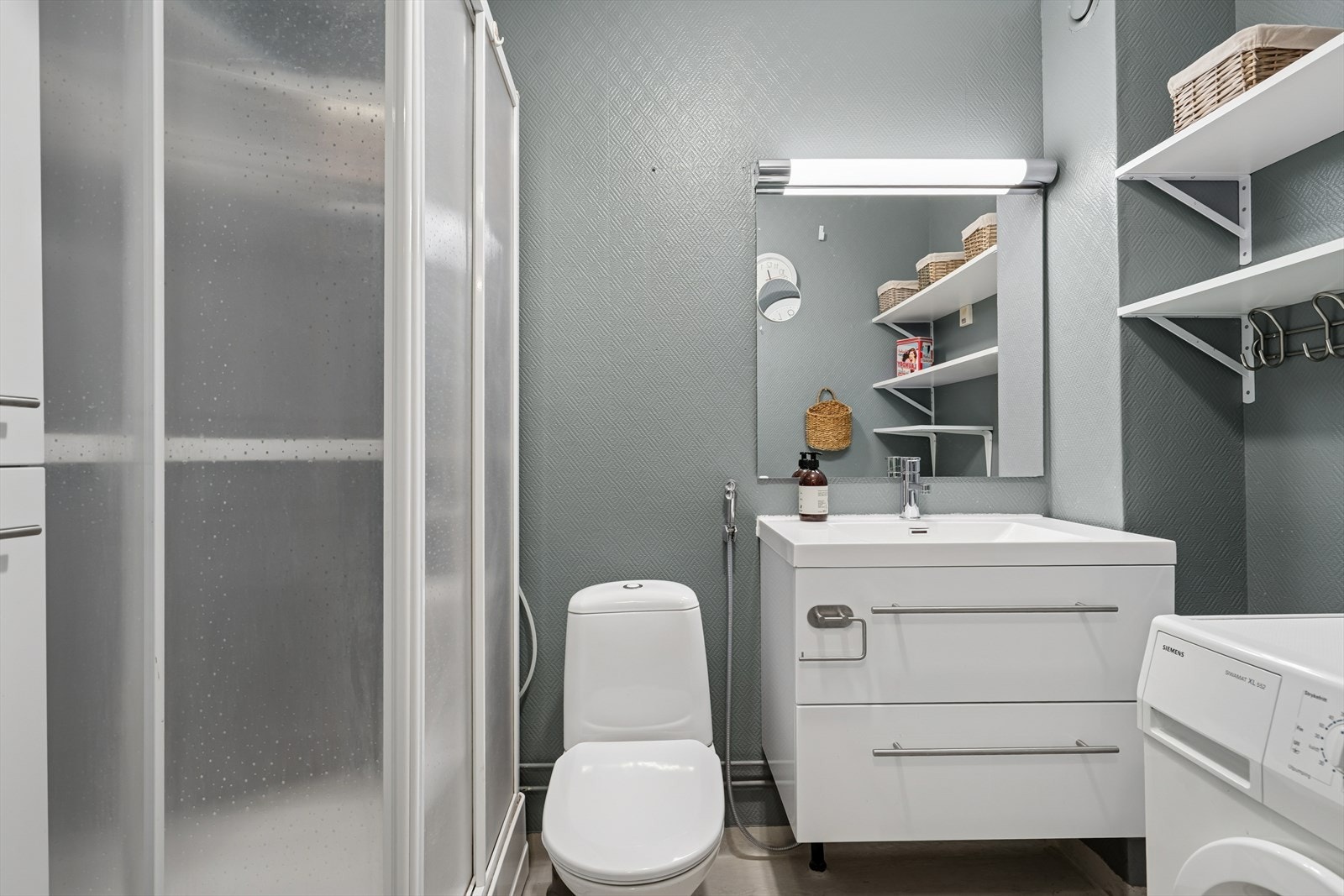 Badet inneholder opplegg for vaskemaskin, innredning med servant fra 2021, veggskap, hyller, dusjkabinett og veggmontert toalett. Galleribilde