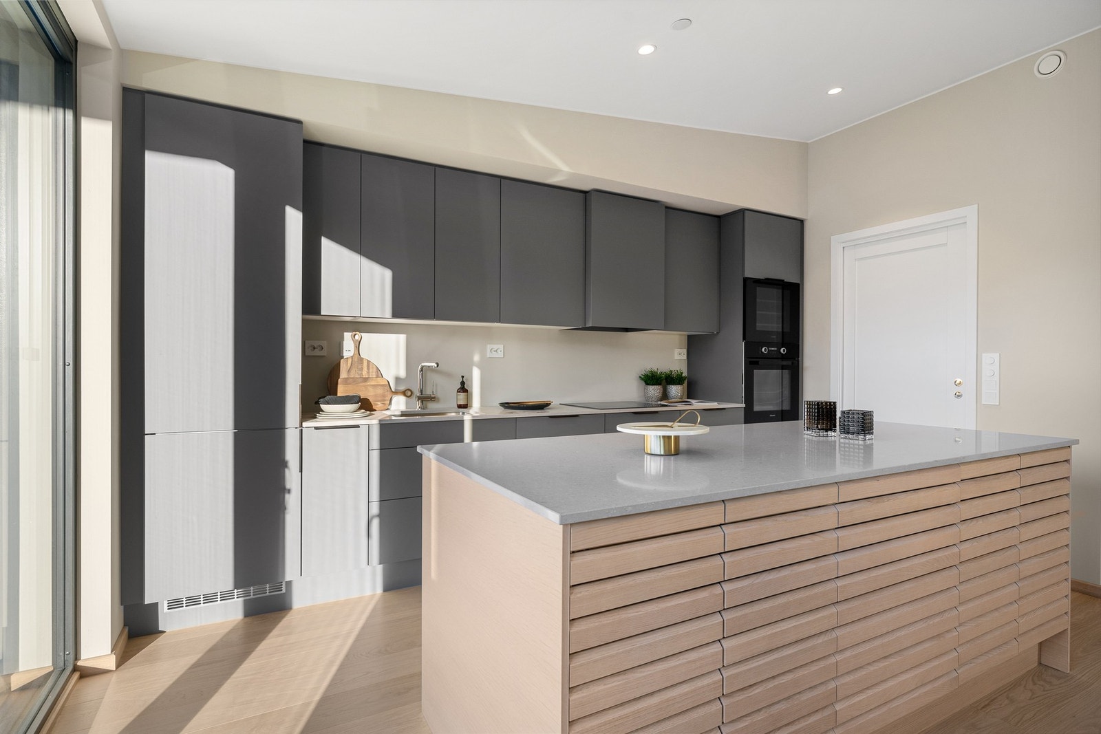 Stilrent kjøkken fra HTH med kompositt benkeplate, romslig kjøkkenøy og integrerte hvitevarer som stekeovn, induksjonstopp, oppvaskmaskin, vinskap og kombinert kjøle-/fryseskap. Galleribilde