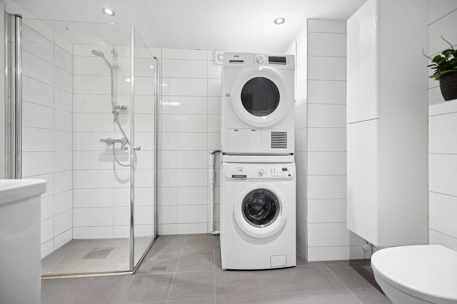 Badet har dusjhjørne med glassdører, vegghengt servantinnredning og veggskap samt vegghengt toalett. Opplegg for vaskemaskin og tørketrommel. Galleribilde