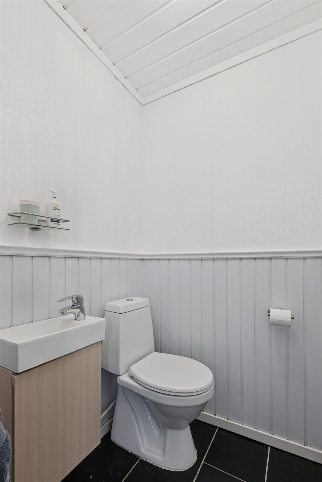 Toalettrommet fremstår som praktisk og funksjonelt, med flislagt gulv som gir et solid og lettstelt underlag. Veggene er kledd med en kombinasjon av panel og tapet. Rommet er innredet med skap for oppbevaring, servant med tilhørende armatur og WC. Galleribilde