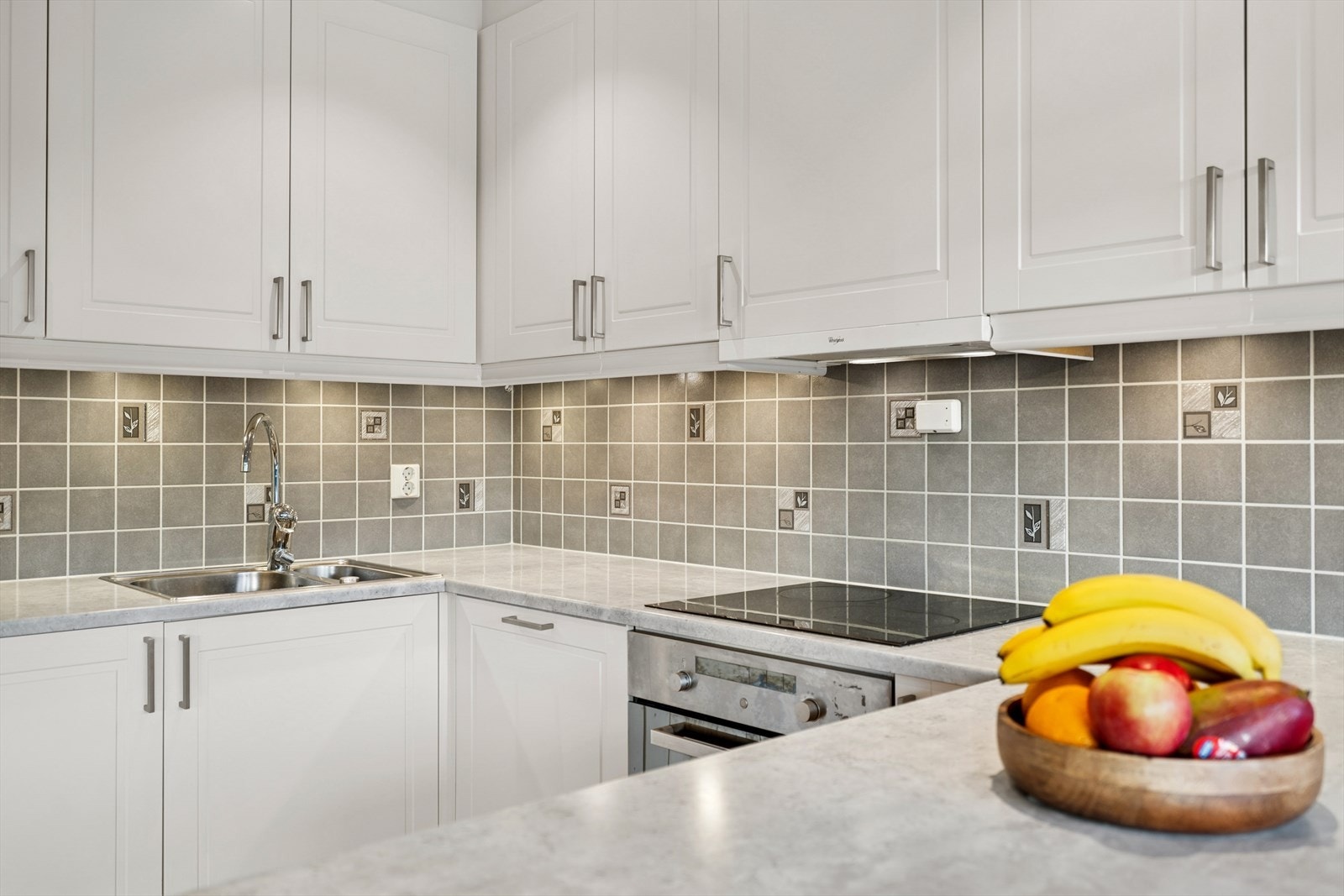 Integrerte hvitevarer av kjøl-/fryseskap, oppvaskmaskin, komfyr, induksjonstopp og avtrekksvifte. Det er fliser mellom benk og overskap som beskytter veggen mot vann og matsøl. Galleribilde