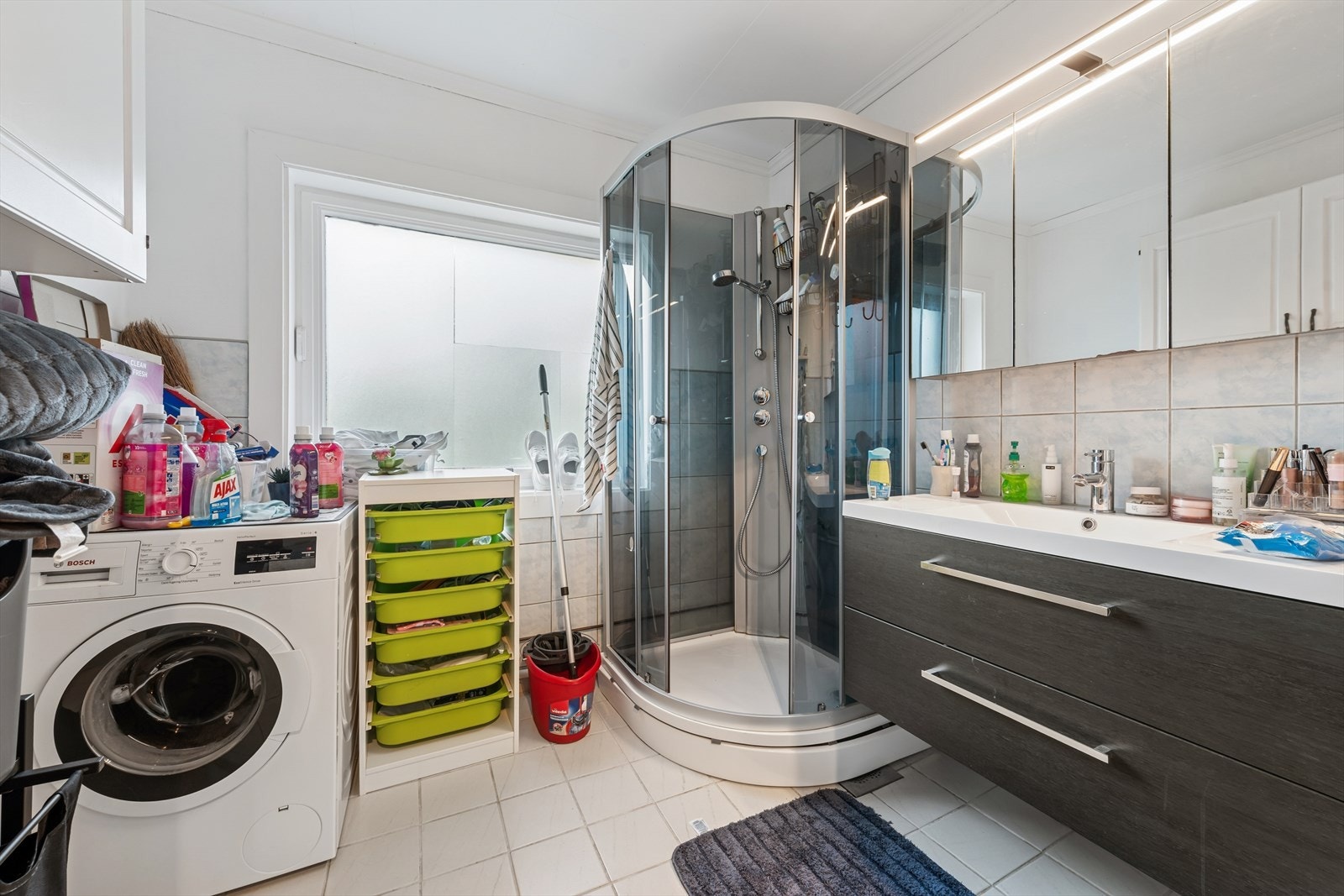 Badet er av eldre dato og har delvis flislagte vegger, takess i himlingen og fliser på betonggulvet. Rommet er innredet med en servant med slette fronter, et dusjkabinett og har opplegg for vaskemaskin. Ventilasjonen på rommet er naturlig. Galleribilde