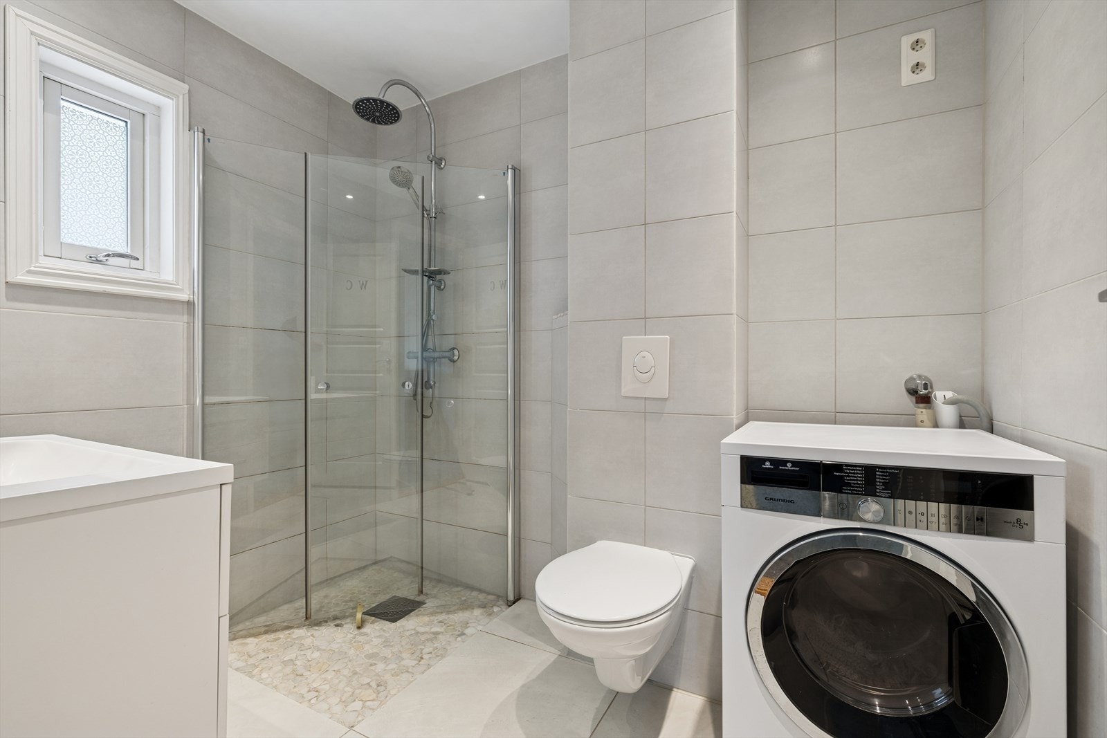 Dusjhjørne med foldedører, vegghengt klosett og servantskap samt speilskap på vegg. Badet har moderne uttrykk og praktiske løsninger, samt opplegg for vaskemaskin. Galleribilde