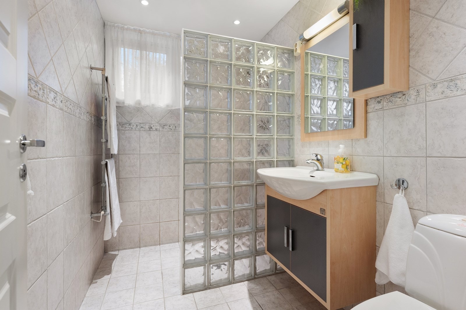 Delikat og pent bad med flislagte vegger og gulv. Badet er innredet med servant, speil med belysning, skap til oppbevaring, dusj, og toalett Galleribilde