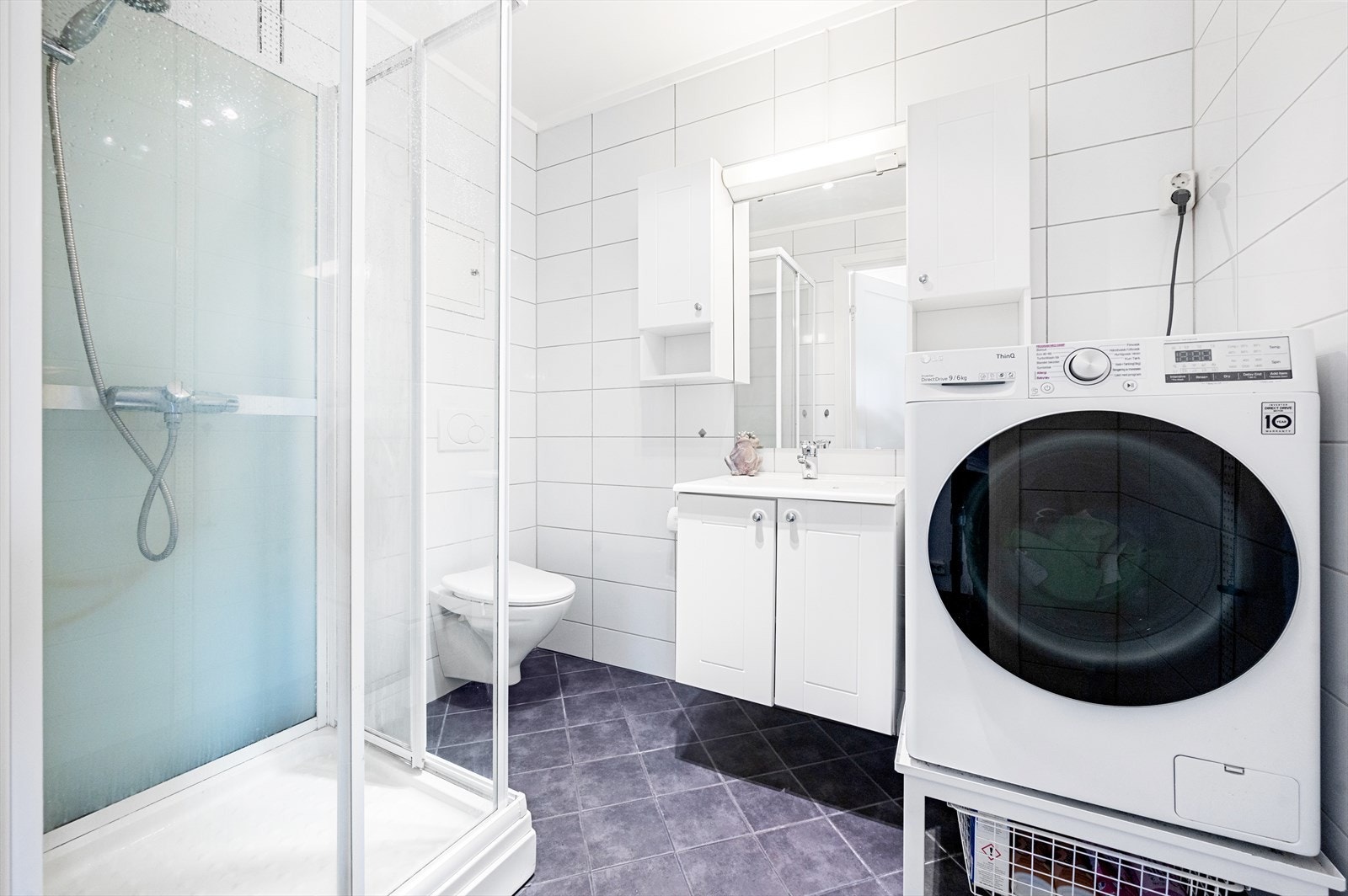 Flislagt bad med varmekabler i gulv, dusjkabinett, vegghengt WC og servant med skap og opplegg til vaskemaskin. Galleribilde