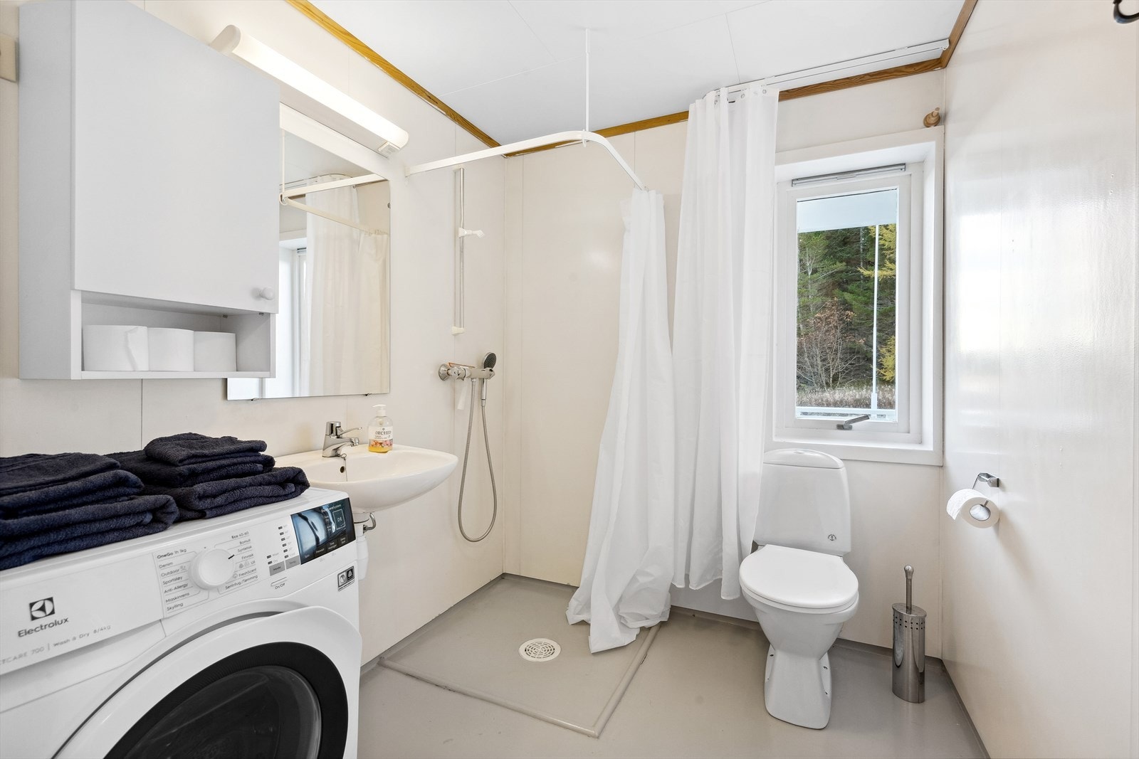 Badet er utstyrt med enkel vegghengt vask, speil med lys, overskap, toalett, dusj med forheng og opplegg for vaskemaskin Galleribilde