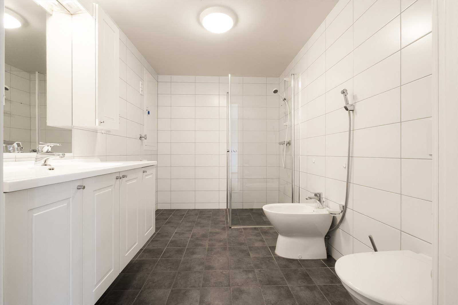 Badet er utstyrt med bidé, vegghengt wc, opplegg for vaskemaskin, og hvit baderomsinnredning med både over- og underskap. Galleribilde