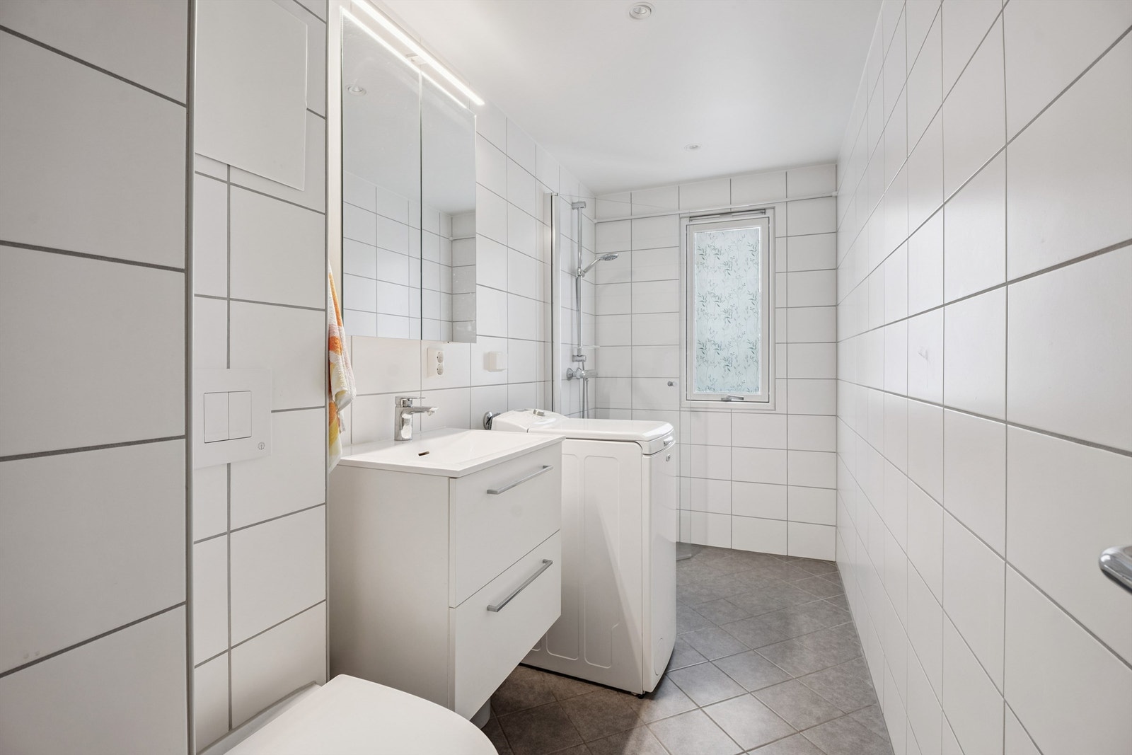 Badet er utstyrt med veggmontert toalett, servantinnredning og opplegg for vaskemaskin. Galleribilde