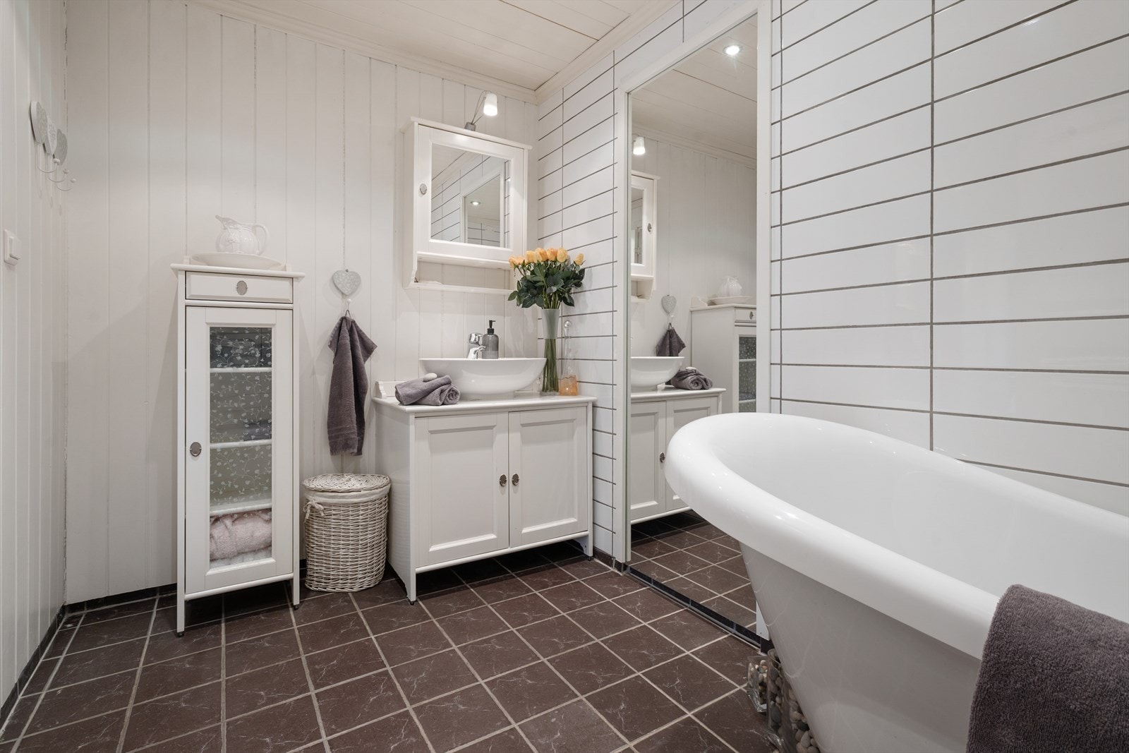 Bad med fliser på gulv og malt trepanel på vegg. Badet har badekar, veggehengt WC og servant på innredning. Galleribilde