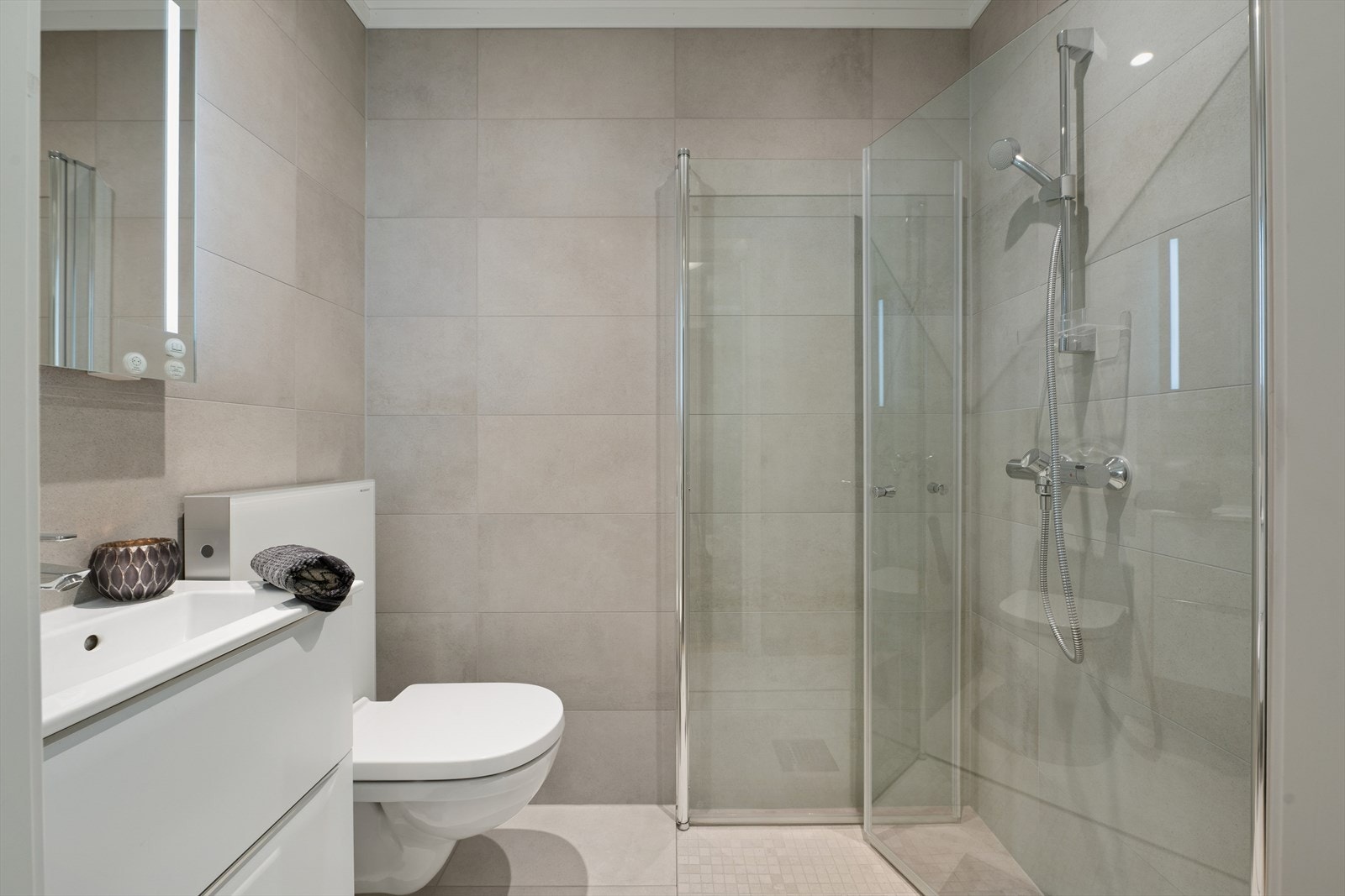 Badet i 1. etasje har flislagte overflater og er innredet med en baderomsinnredning med heldekkende servant, speil med belysning, veggmontert toalett og dusj med innfellbare dusjdører. Galleribilde