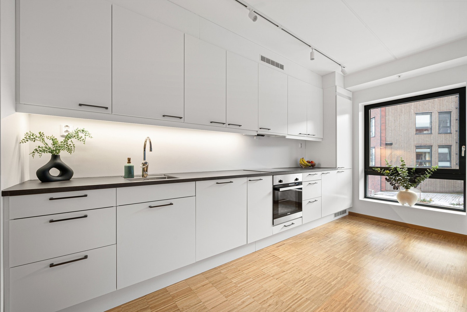 Kjøkkenet fremstår som moderne og stilrent, med lyse fronter og mørk benkeplate. Over sonen er det installert en praktisk spotskinne. Galleribilde