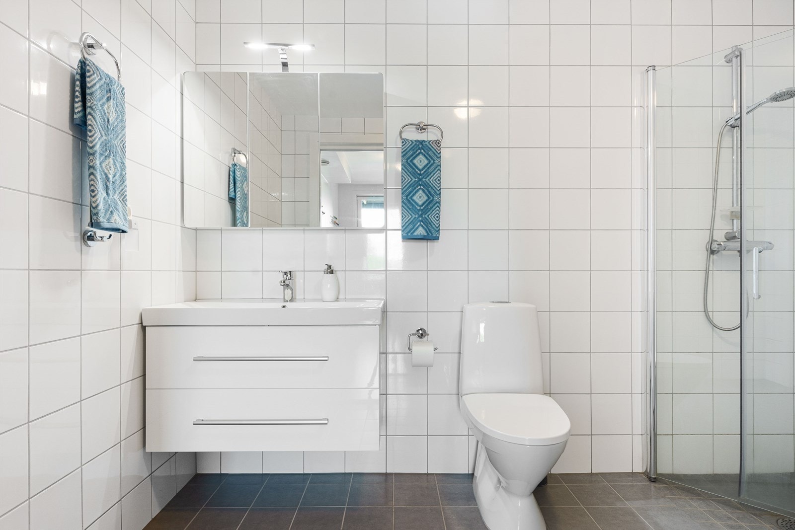 Romslig og delikat baderom med dusjhjørne, servant i innredning med skuffer og speil overskap. Wc og opplegg for vaskemaskin og tørketrommel.
Badet har fliser på vegger, gulv og malt i himlinger. Galleribilde