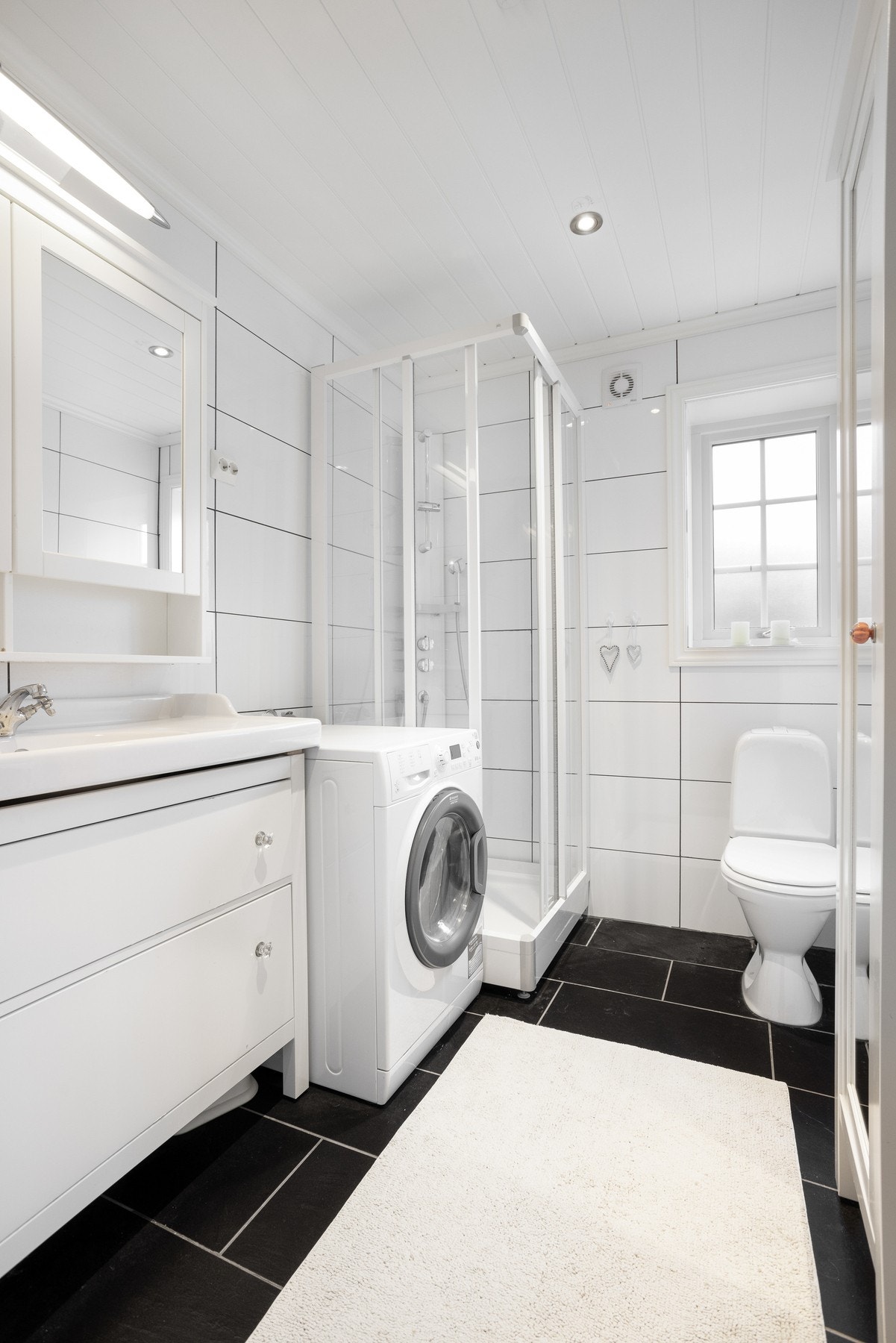 Flislagt baderom med toalett, dusjkabinett, innredning med servant og opplegg til vaskemaskin Galleribilde