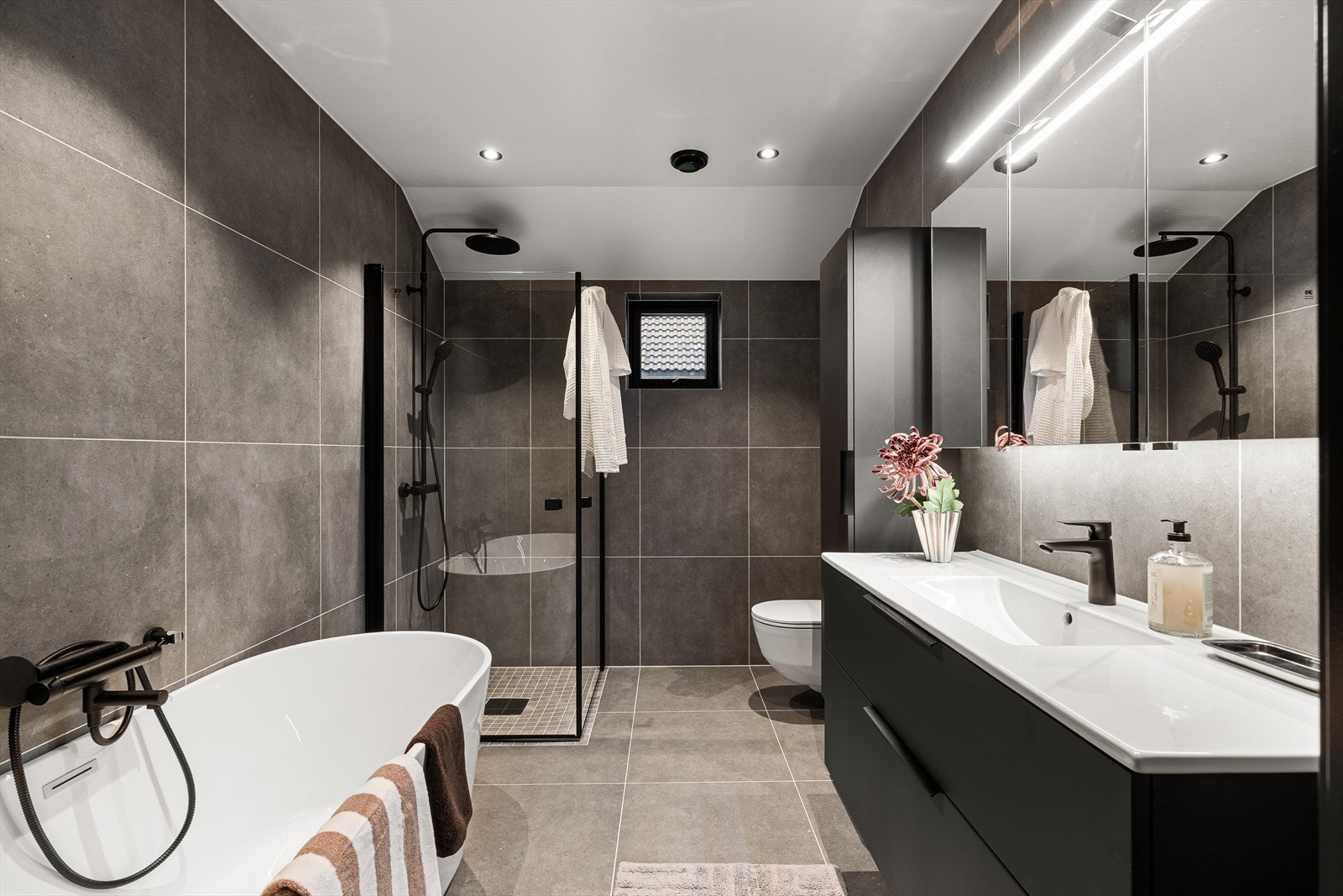 Delikat baderom med fliser på gulv og vegger. Badet er innredet med vask i seksjon, speil med belysning, frittstående badekar, vegghengt toalett og dusjhjørne. Galleribilde