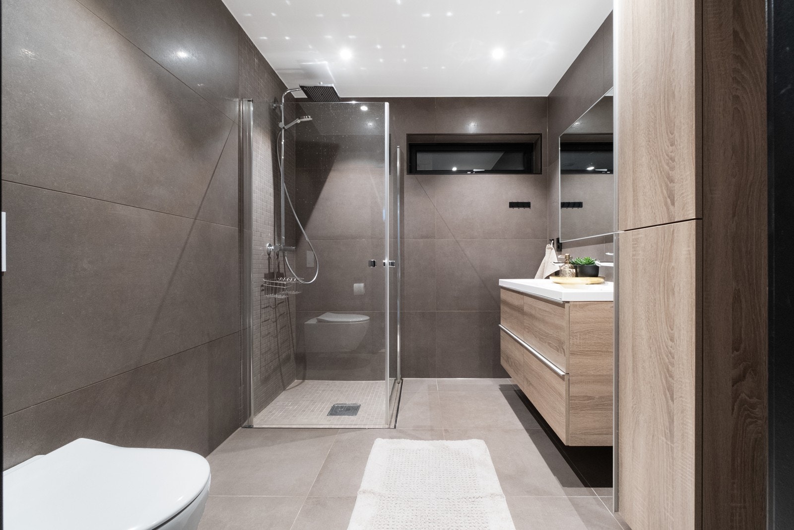 Flislagt bad utstyrt med dusjhjørne med tropedusj og glassvegger, vegghengt toalett og baderomsinnredning med heldekkende servant. Galleribilde