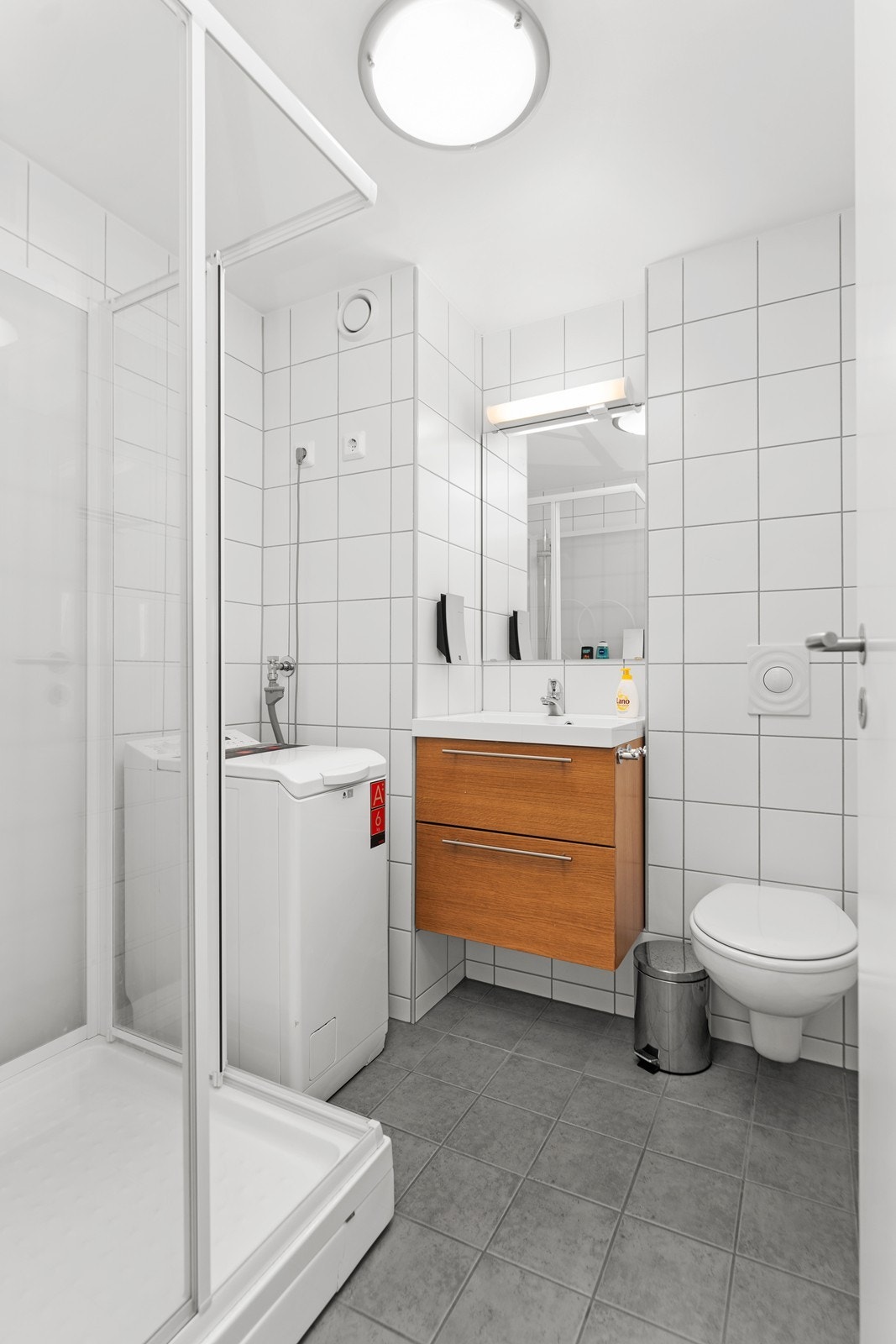 Bad i leiligheten med dusjhjørne, vegghengt toalett, servant og opplegg til vaskemaskin Galleribilde