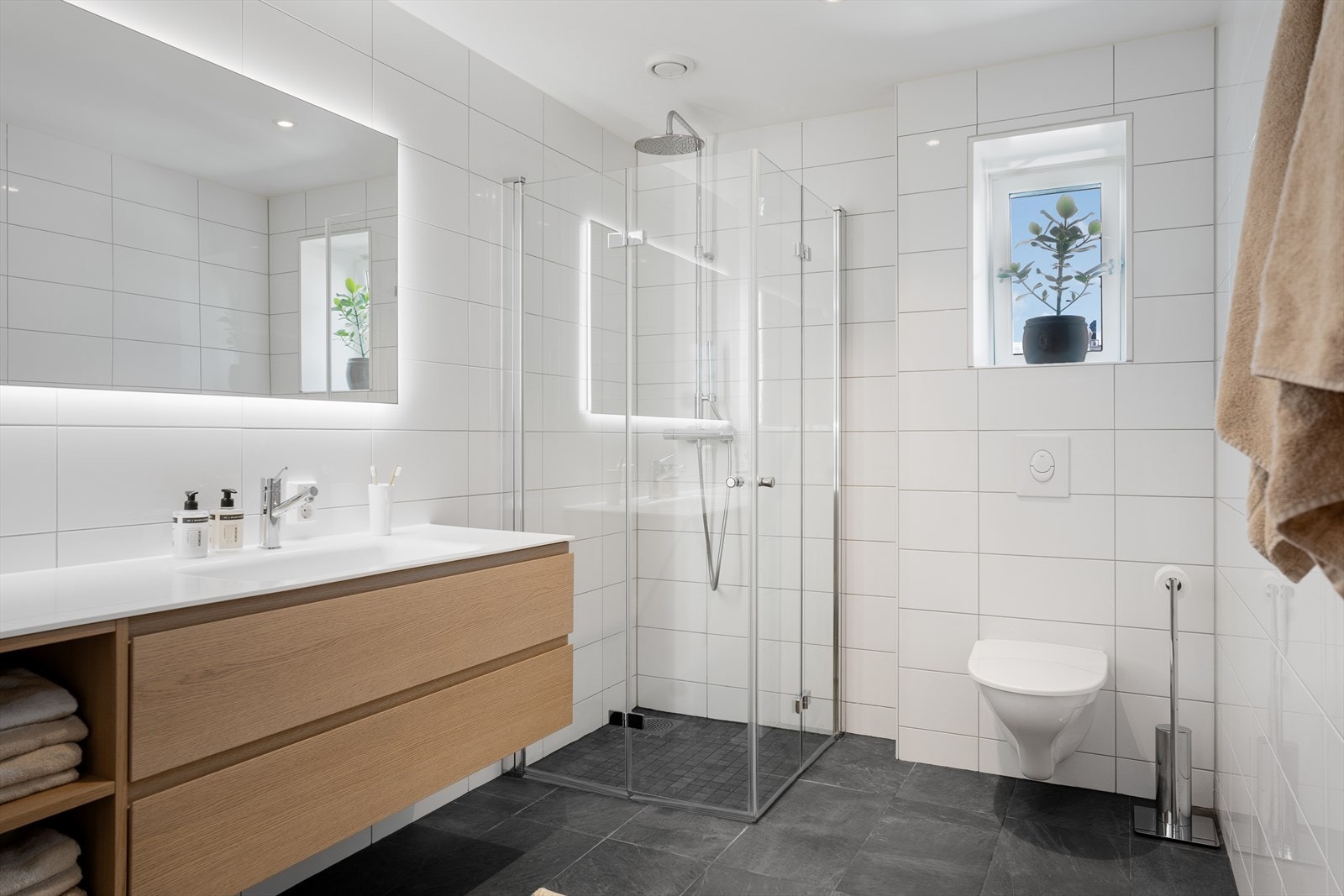 Stort, lekkert og helfliset bad som inneholder baderomsinnredning med heldekkende servant, dusjhjørne og vegghengt toalett. Galleribilde