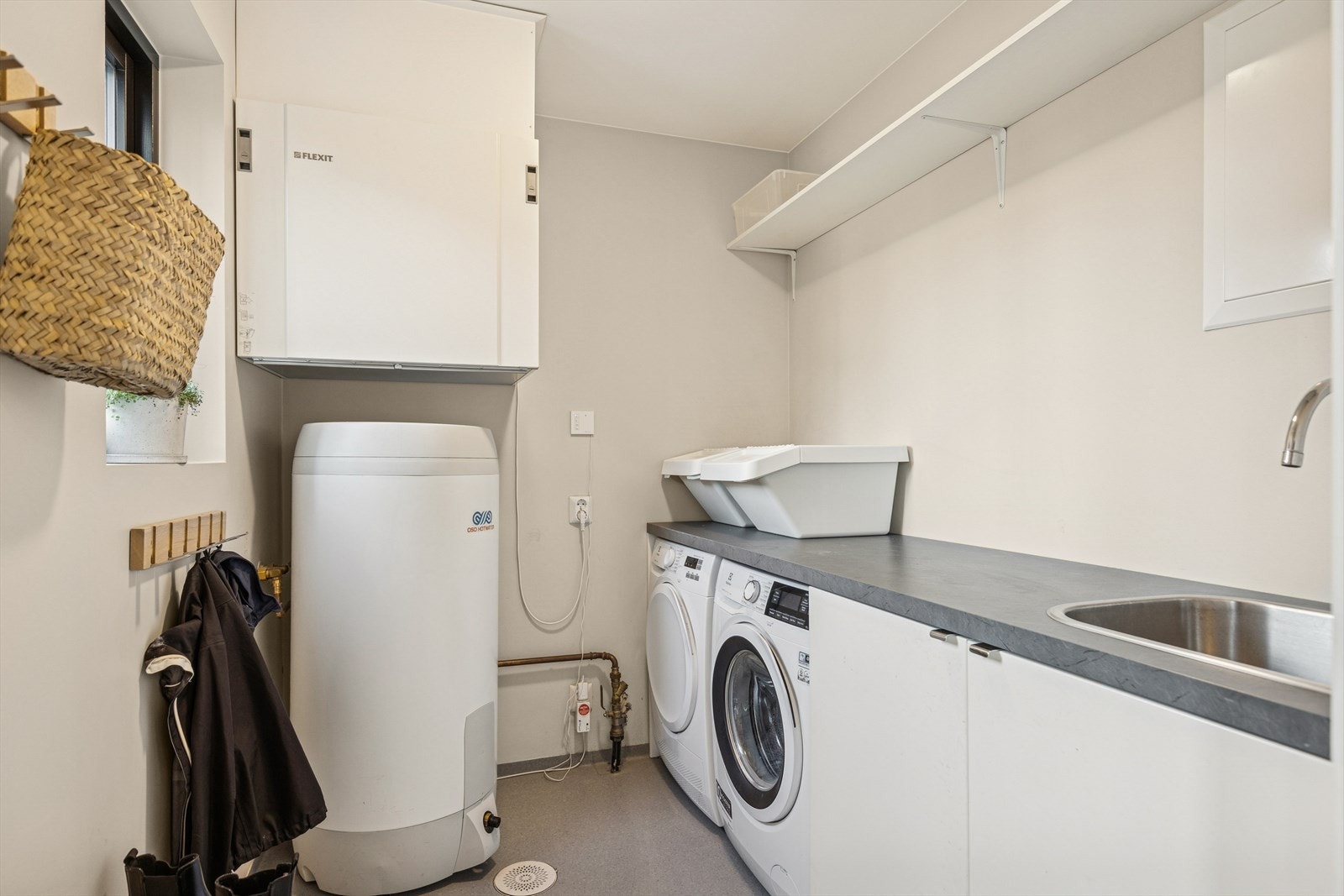 Praktisk vaskerom med gulvbelegg på gulv. Rommet har utslagsvask, opplegg for vaskemaskin, fordelerskap for vannrør og varmtvannsbereder. Galleribilde