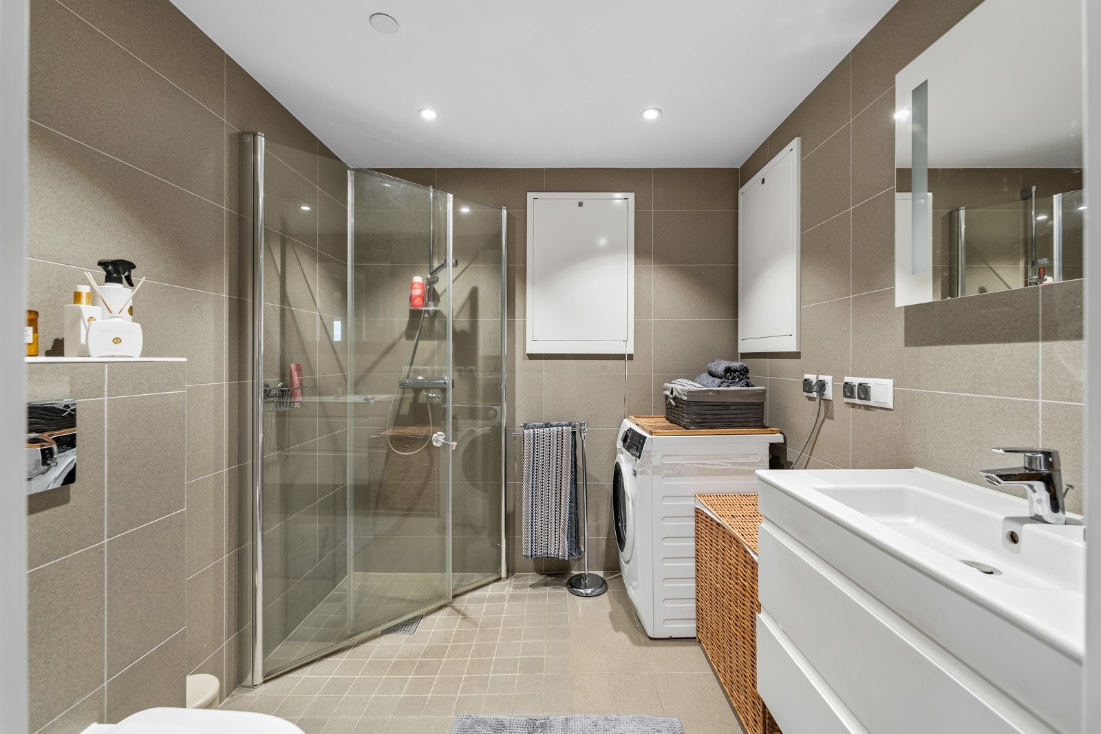 Badene er pent innredet med heldekkende servant på hvit skuffeseksjon, veggmontert toalett, speil med integrert belysning og et dusjhjørne med innfellbare glassdører. Det er også opplegg for vaskemaskin på begge bad Galleribilde