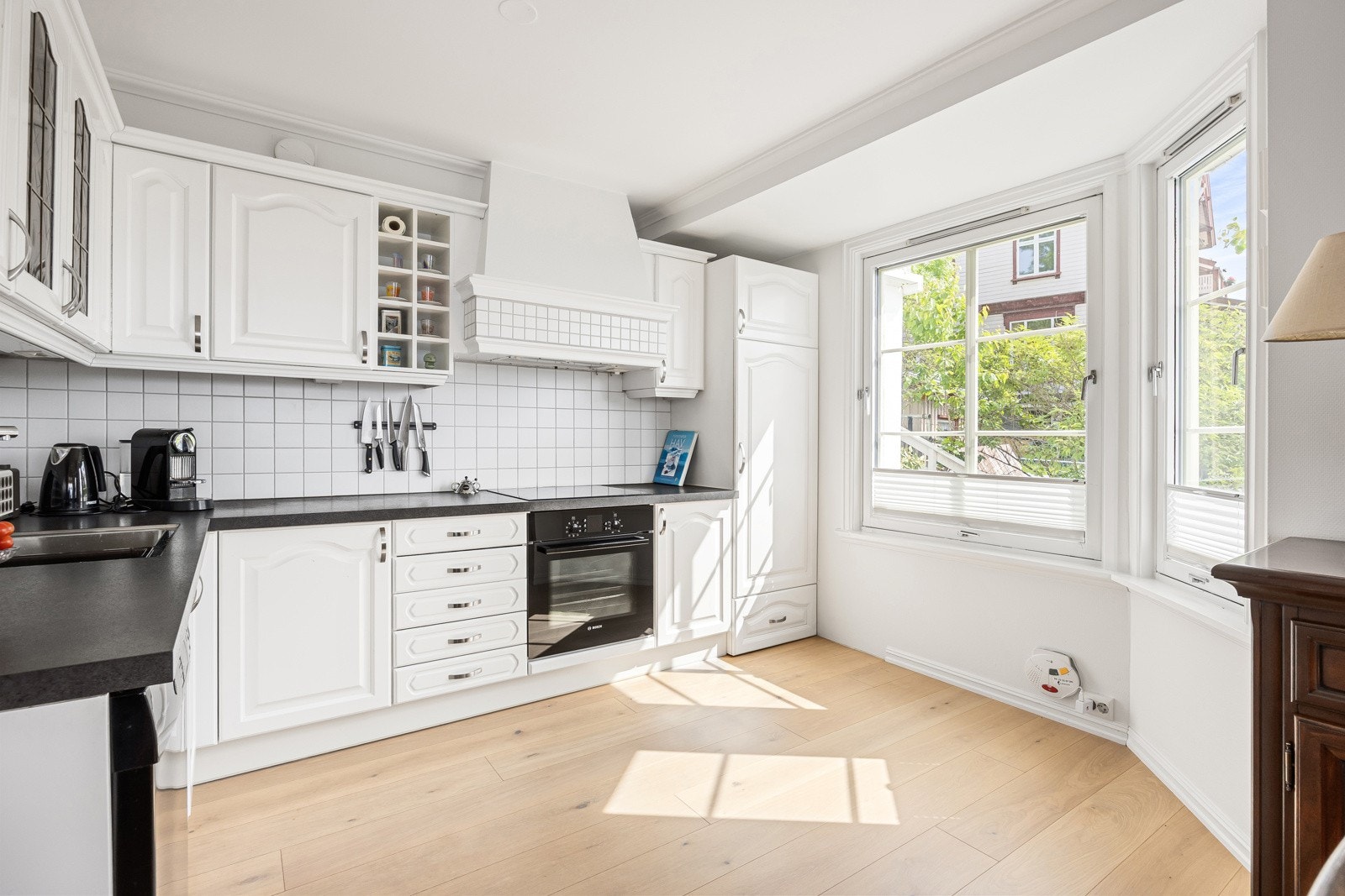 Kjøkkeninnredningen er plassert i L-form, med hvite, profilerte fronter, vitrineskap og plassbygd ventilatorhette. Galleribilde