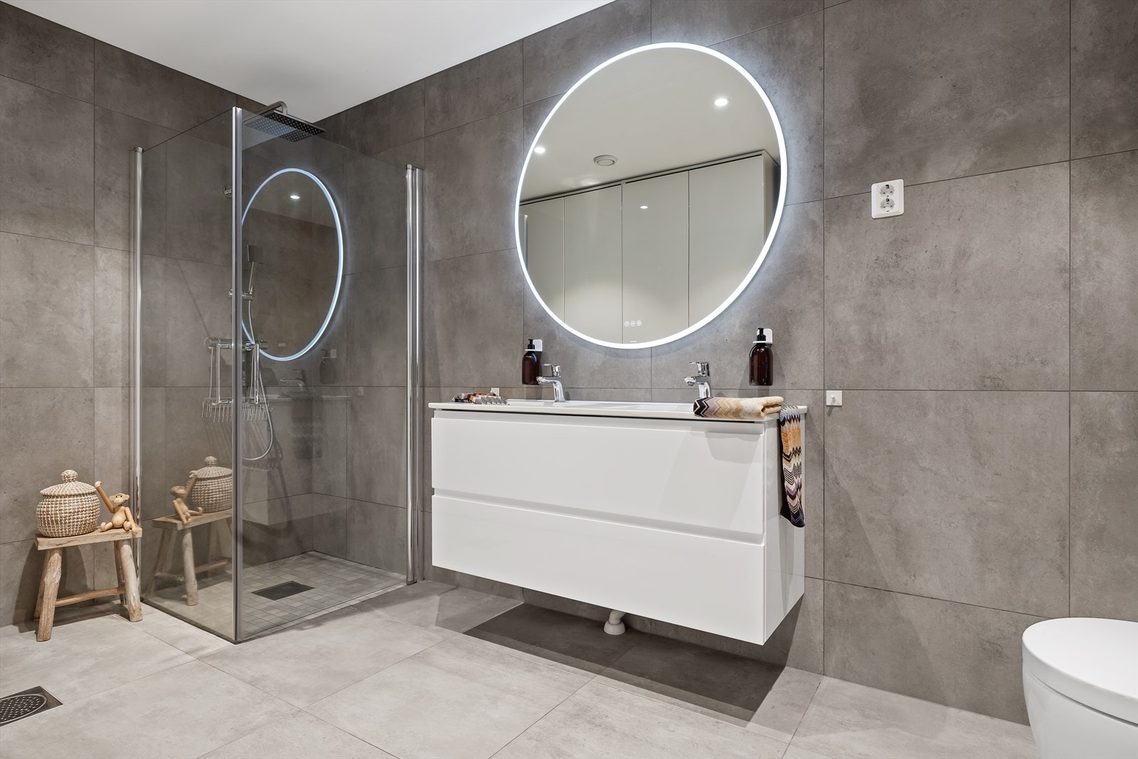 Badet innehar dusjhjørne med innfellbare glassvegger, vegghengt toalett og dobbel servantinnredning med skuffer. Galleribilde