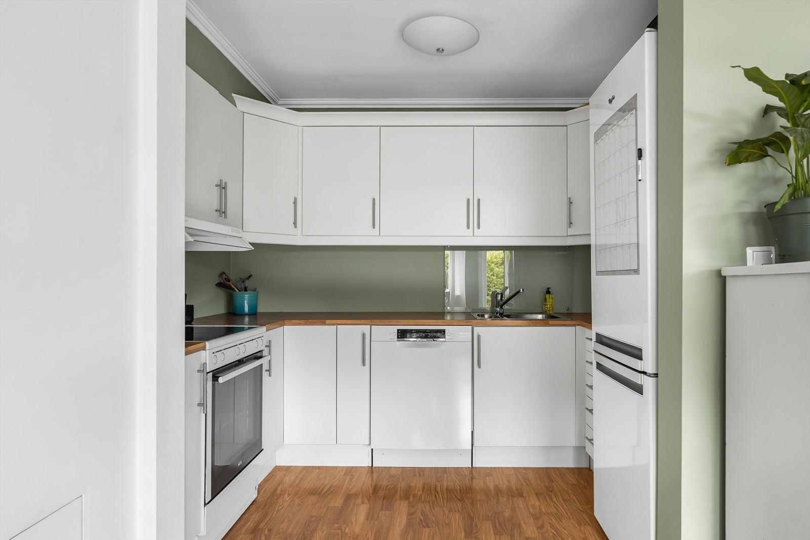 Kjøkken med lys innredning innredet med laminerte skrog, glatte fronter og laminert benkeplate med kjøkkenkum. Laminatplater og belysning over benkeplate. Plass til frittstående komfyr, oppvaskmaskin og kombiskap kjøl/frys. Galleribilde