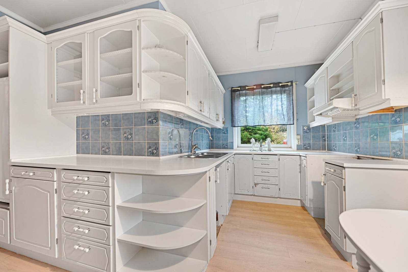 Kjøkkenet, en gang hjertet av hjemmet, ser utdatert ut med laminatgulv, uten integrerte hvitevarer og med slitte skap med profilerte fronter. Galleribilde