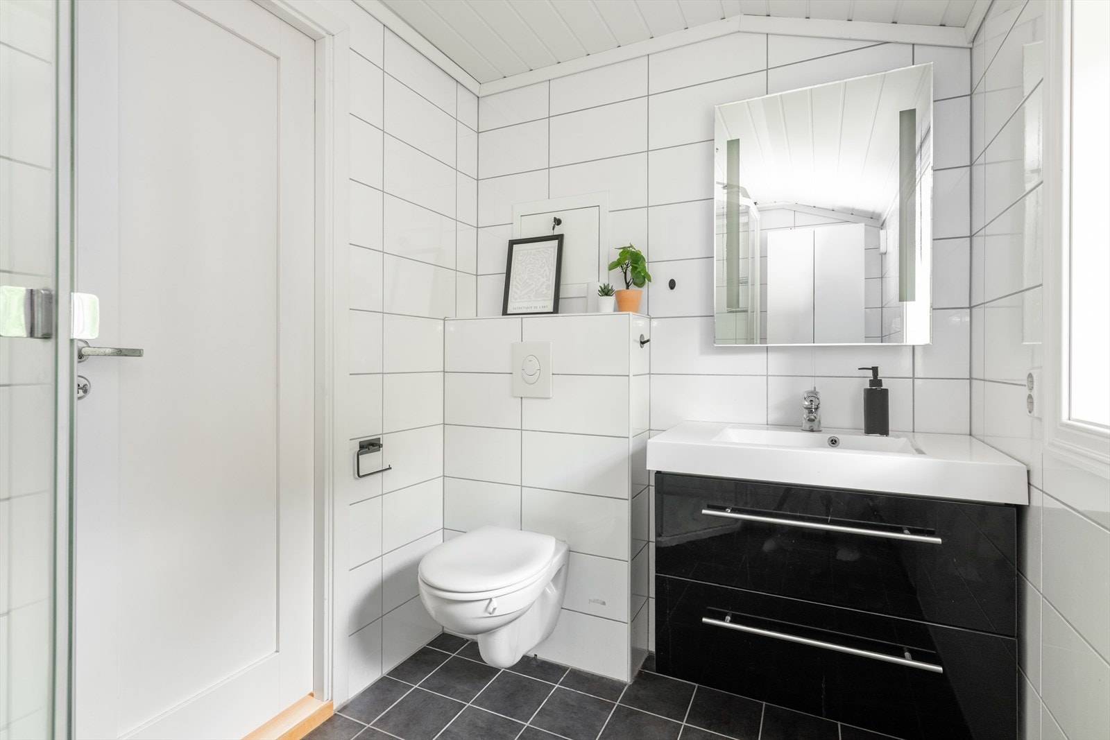 Badet inneholder veggfestet toalett, dusjkabinett og servantinnredning med underskuffer, speil med innebygget LED-lys, samt veggfeste skap. Galleribilde