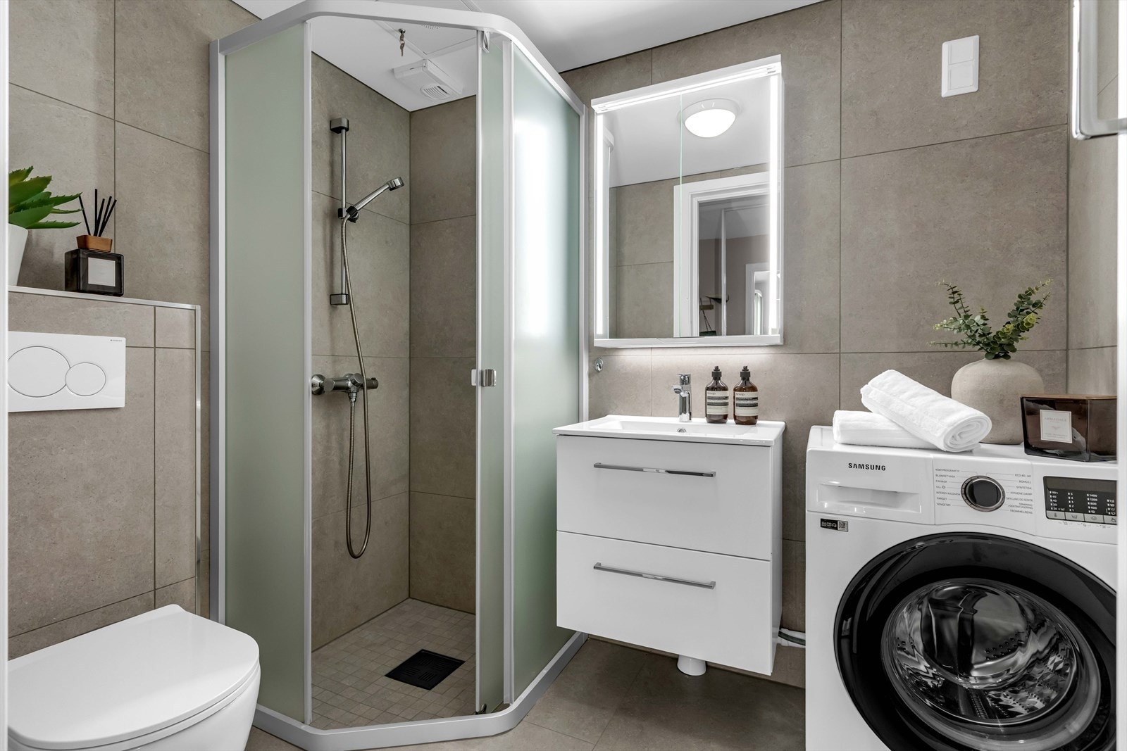 Flislagt gulv og vegger i sandtone. Badet er bygget opp med vegghengt toalett, dusjnisje og vegghengt servantinnredning. Galleribilde