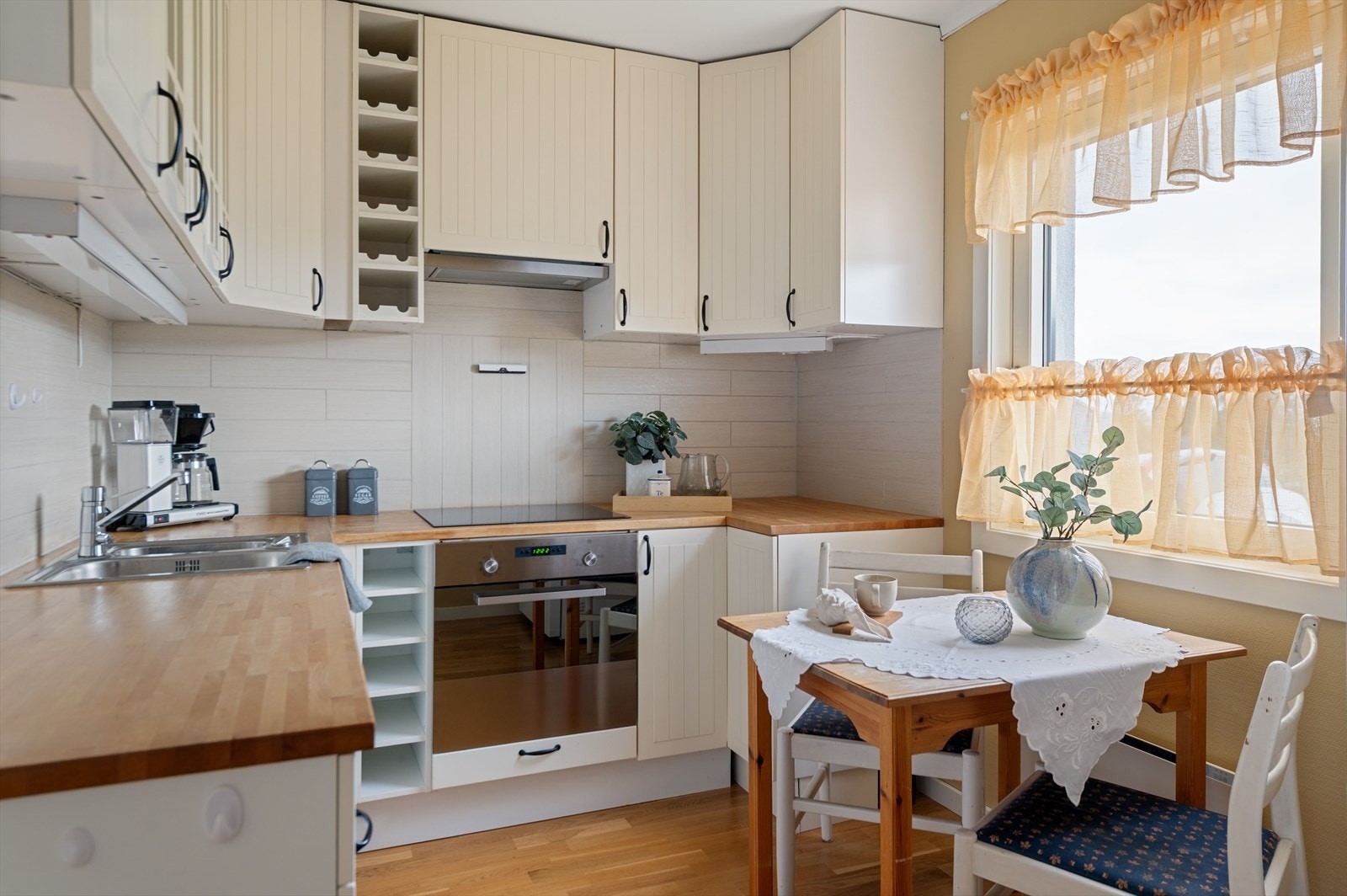 Lys kjøkkeninnredning innredet med laminerte skrog, profilerte fronter og heltre benkeplate med kjøkkenkum. Integrert koketopp og komfyr, med plass for frittstående oppvaskmaskin og kjøleskap. Galleribilde