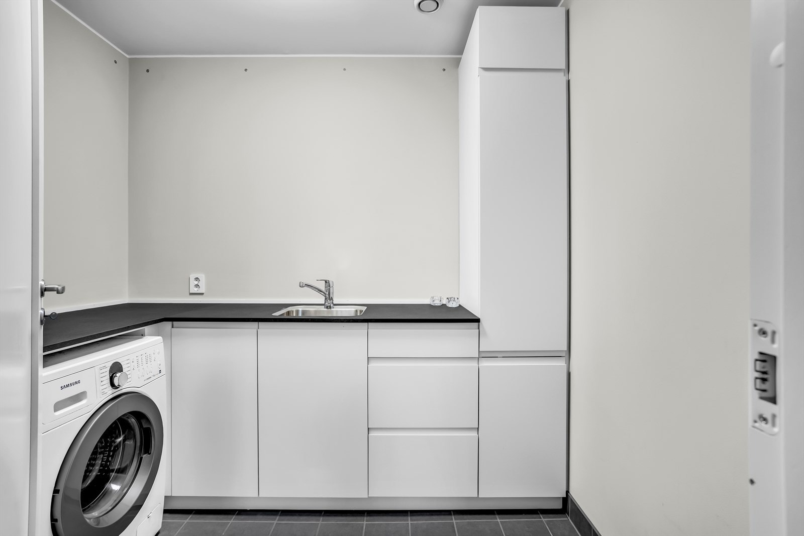 Vaskerommet er utstyrt med hvit skapinnredning, laminat benkeplate, nedfelt frustfri stålkum og opplegg for vaskemaskin. Galleribilde