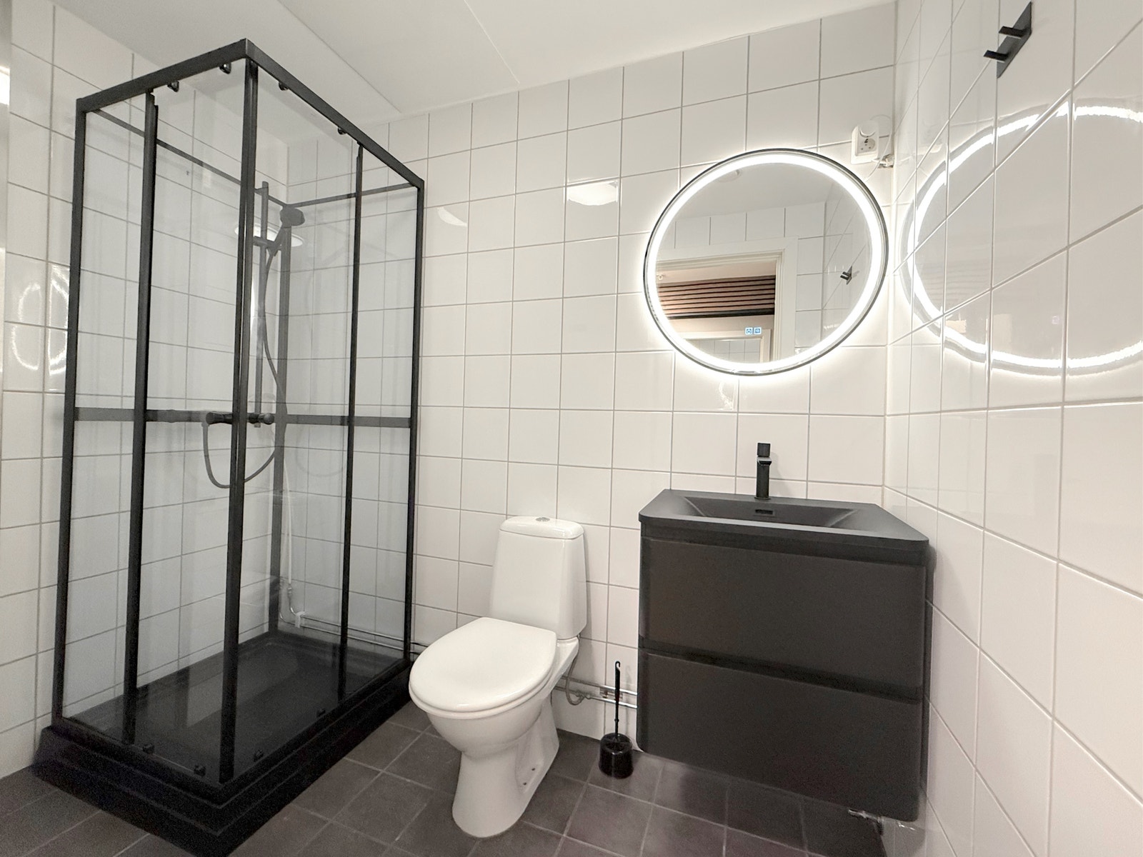 Flislagt bad med sort dusjkabinett, toalett, servant i mørk innredning og rundt speil med lys Galleribilde