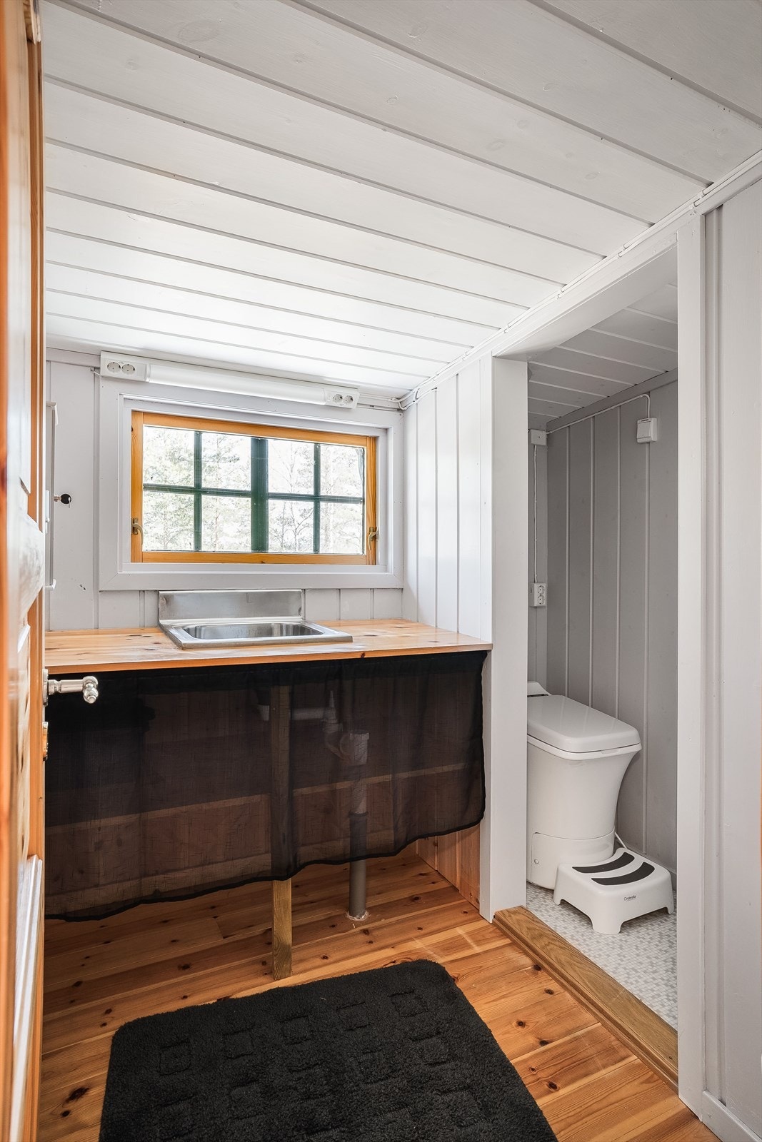 Rett innenfor døren ligger sanitærrom med Cinderella forbrenningstoalett og hyttedusj. Rommet er malt i en moderne gråfarge. Innredning med nedfelt servant på benkeplate med hyller under. Vindu og belysning over vask. Galleribilde