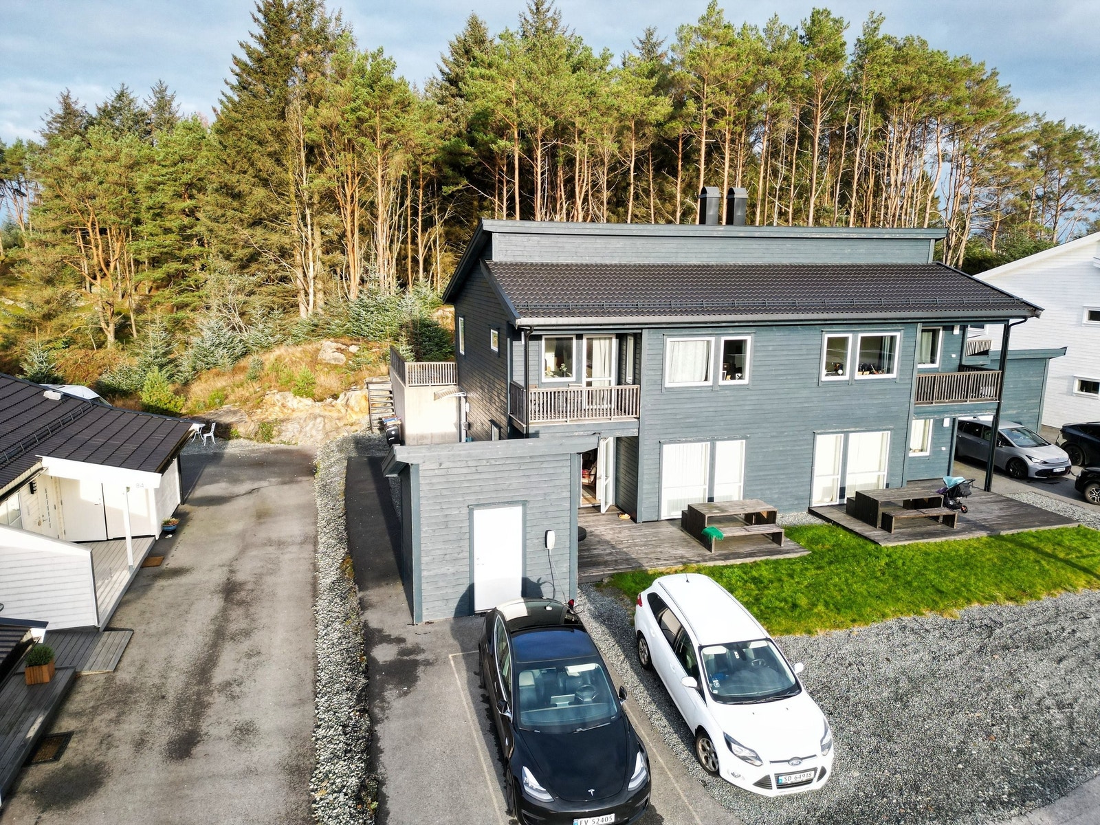 Oversiktsbilete av bustaden med solrik terrasse, parkeringsplassar rett utanfor og naturen som næraste nabo. Den lune og grøne skogkanten bak huset bidreg til eit roleg og triveleg bumiljø. Galleribilde
