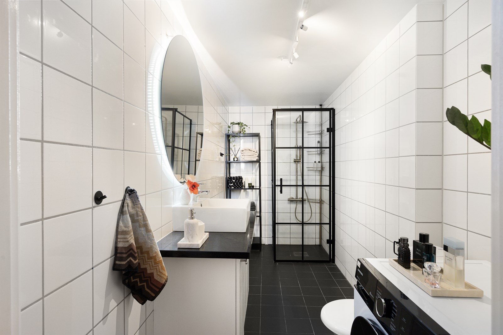 Flislagt baderom innredet med klosett, nedfelt servant i innredning, speil med belysning og regnfalldusj med svingdører. Galleribilde