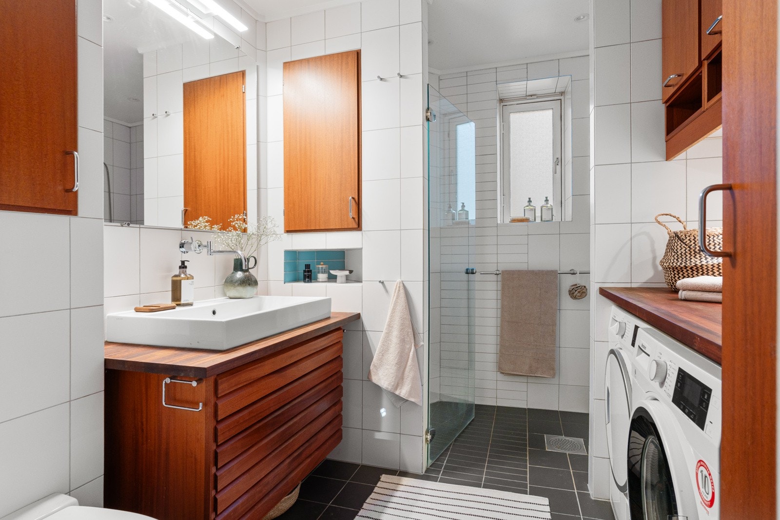 Flislagt bad med varmekabler, dobbel dusjnisje, veggmontert servantinnredning og toalett som opplegg for både vaskemaskin og tørketrommel Galleribilde