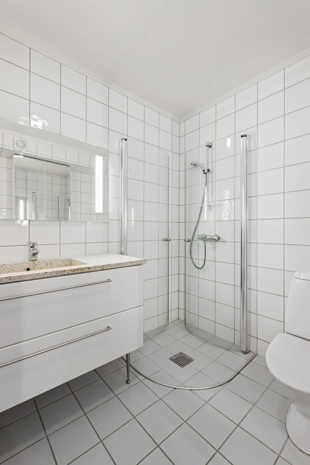 Badet er også innredet med et gulvmontert toalett, veggmontert dusj med buede glassdører og opplegg for vaskemaskin. Galleribilde