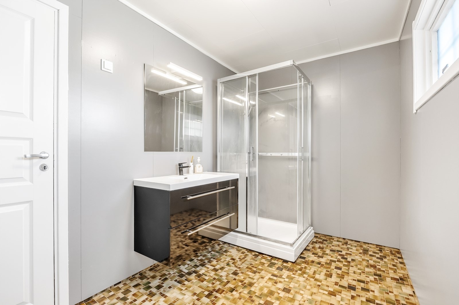 Badet har lysmalte våtromsplater på veggene og fliser på gulvet med lun varme. Innredningen består av heldekkende servant over sort, høyglans skuffeseksjon med stort speil i overkant. Galleribilde