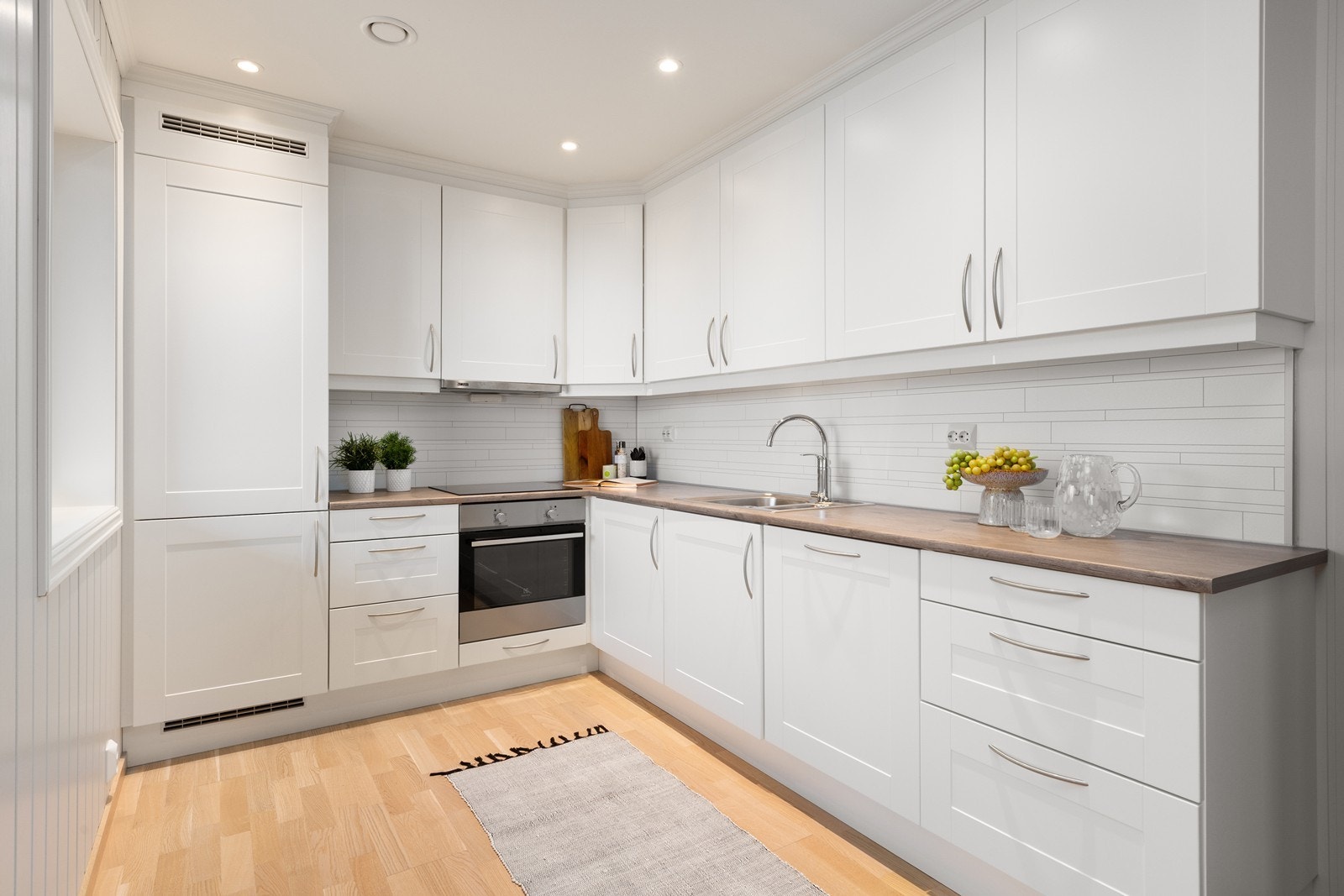 Innredningen har lyse, profilerte fronter og benkeplate i laminat med nedfelt vask, samt integrerte hvitevarer som stekeovn, induksjonstopp, oppvaskmaskin og kjøl-/fryseskap. Galleribilde