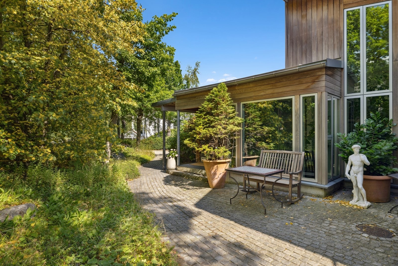 Velstelt og innbydende inngangsparti med store glassflater, grønne omgivelser og hyggelige detaljer som benk, beplantning og skulptur. Galleribilde