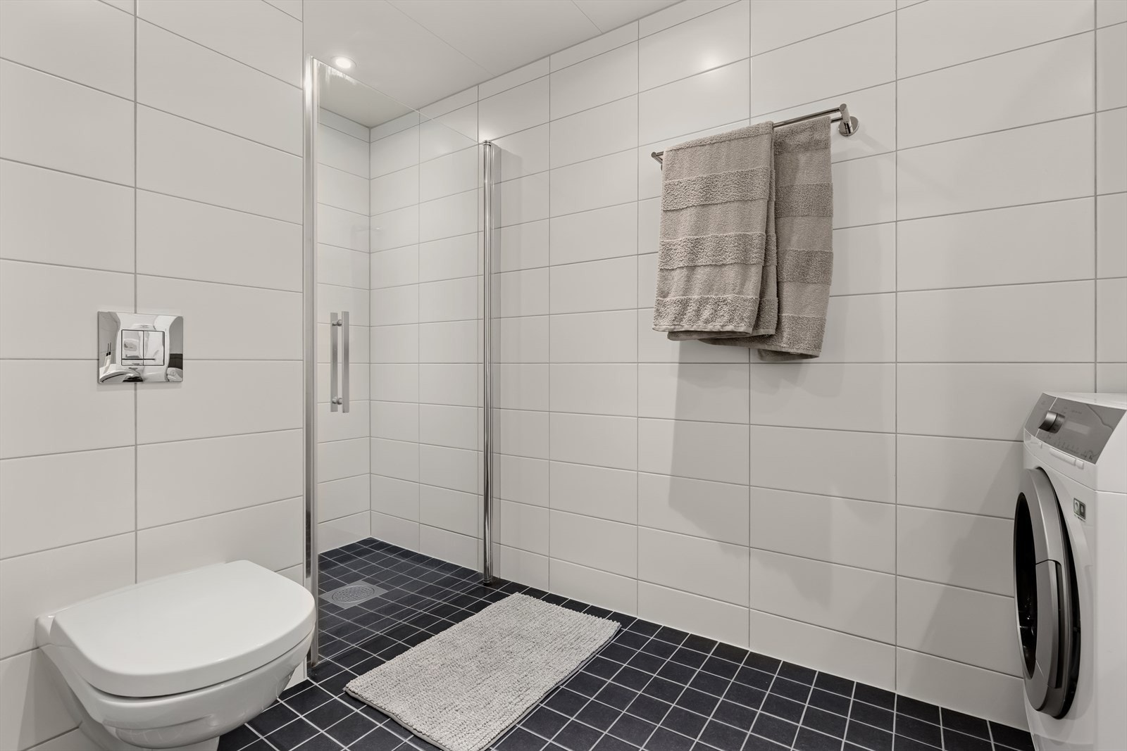 Baderommet har en hvit innredning med toalett, dusjhjørne, servant med skuffer og speil, og opplegg til vaskemaskin Galleribilde