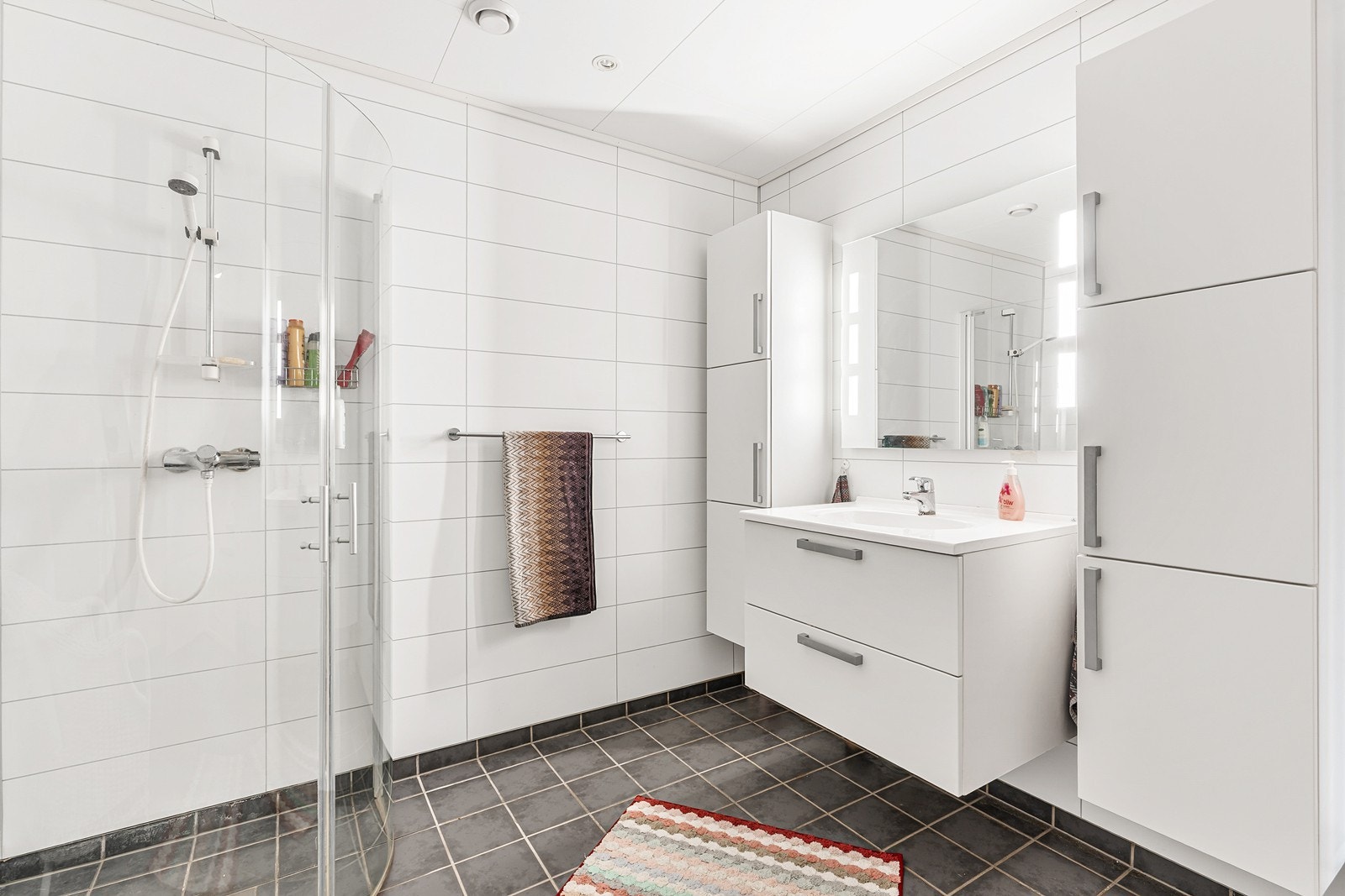 Romslig og tidsriktig bad nnredet med dusjløsning med glassdører, heldekkende vask med skuffeinnredning, speil, to høyskap for ekstra lagring, samt et toalett. Galleribilde