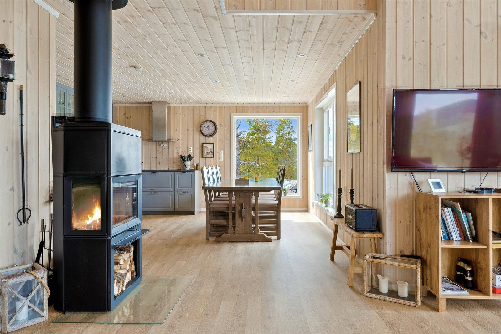 Stue med peisovn, gjennomgående trepanel og åpen løsning mot kjøkken og spiseplass. Galleribilde