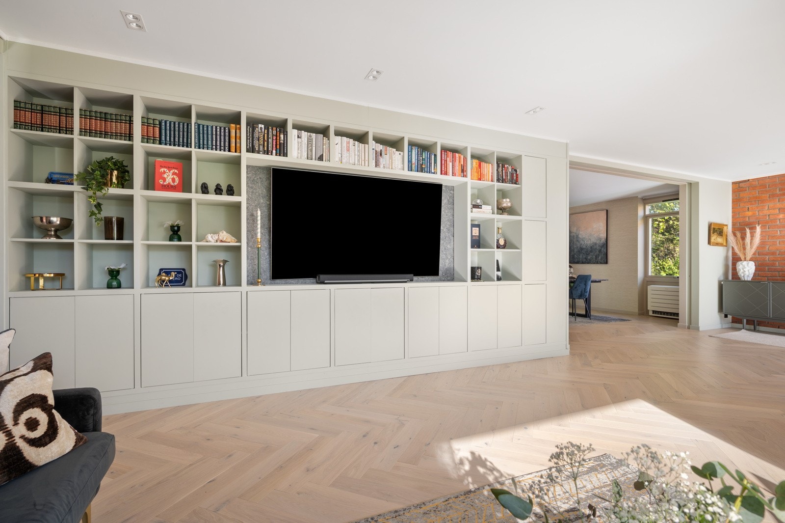 Et innebygget hyllesystem strekker seg over hele stueveggen og gir rikelig med oppbevaringsplass og et gjennomført uttrykk Galleribilde