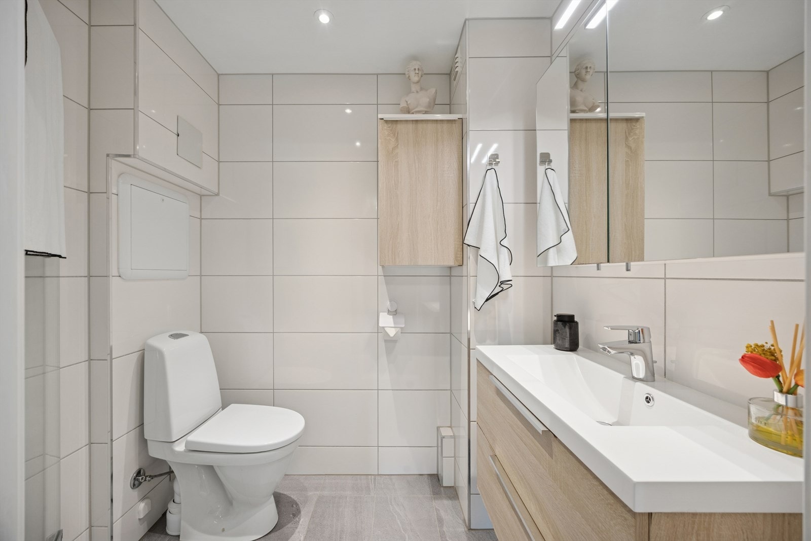 Pent flislagt bad med servant med underskap, overskap, overskap med speildører og lys, dusjhjørne med takdusj og svingbare glassdører og frittstående toalett. Galleribilde