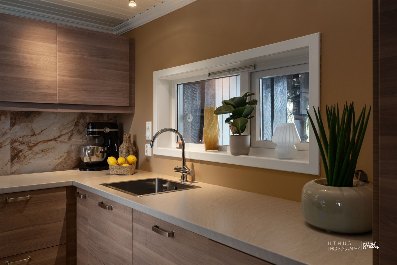 Innredningen består av heltre, glatte skapfronter med over- og underskap som gir god oppbevaringsplass. Benkeplaten er i laminat med lys overflate, og det er montert nedfelt platetopp og rustfri oppvaskkum med avrenningsfelt. Veggen over benk er flislagt. Galleribilde