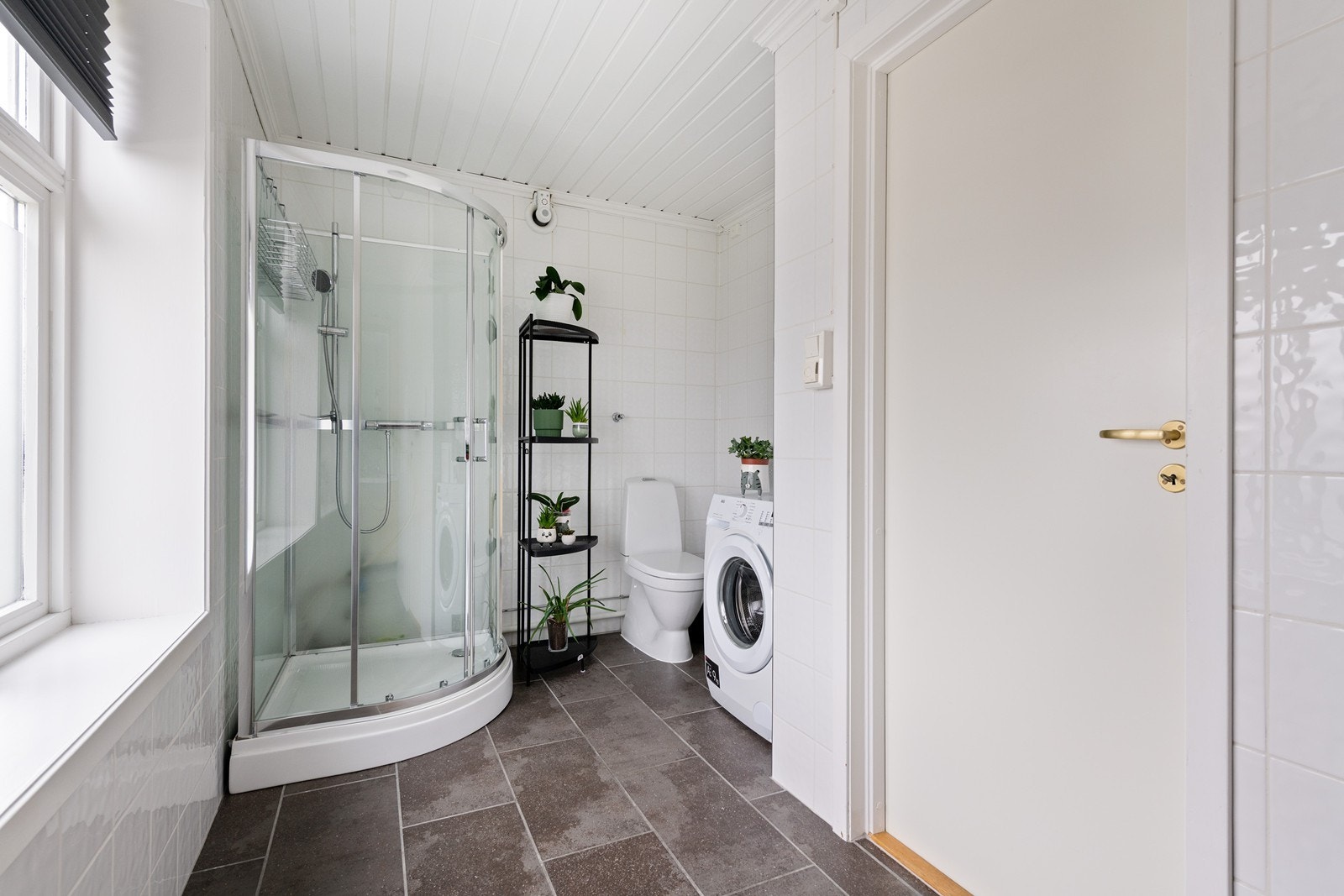 Badet er lyst og med mørke fliser langs gulv. Det er innredet med dusjkabinett, wc, servant med oppbevaring og opplegg for vaskemaskin. Galleribilde